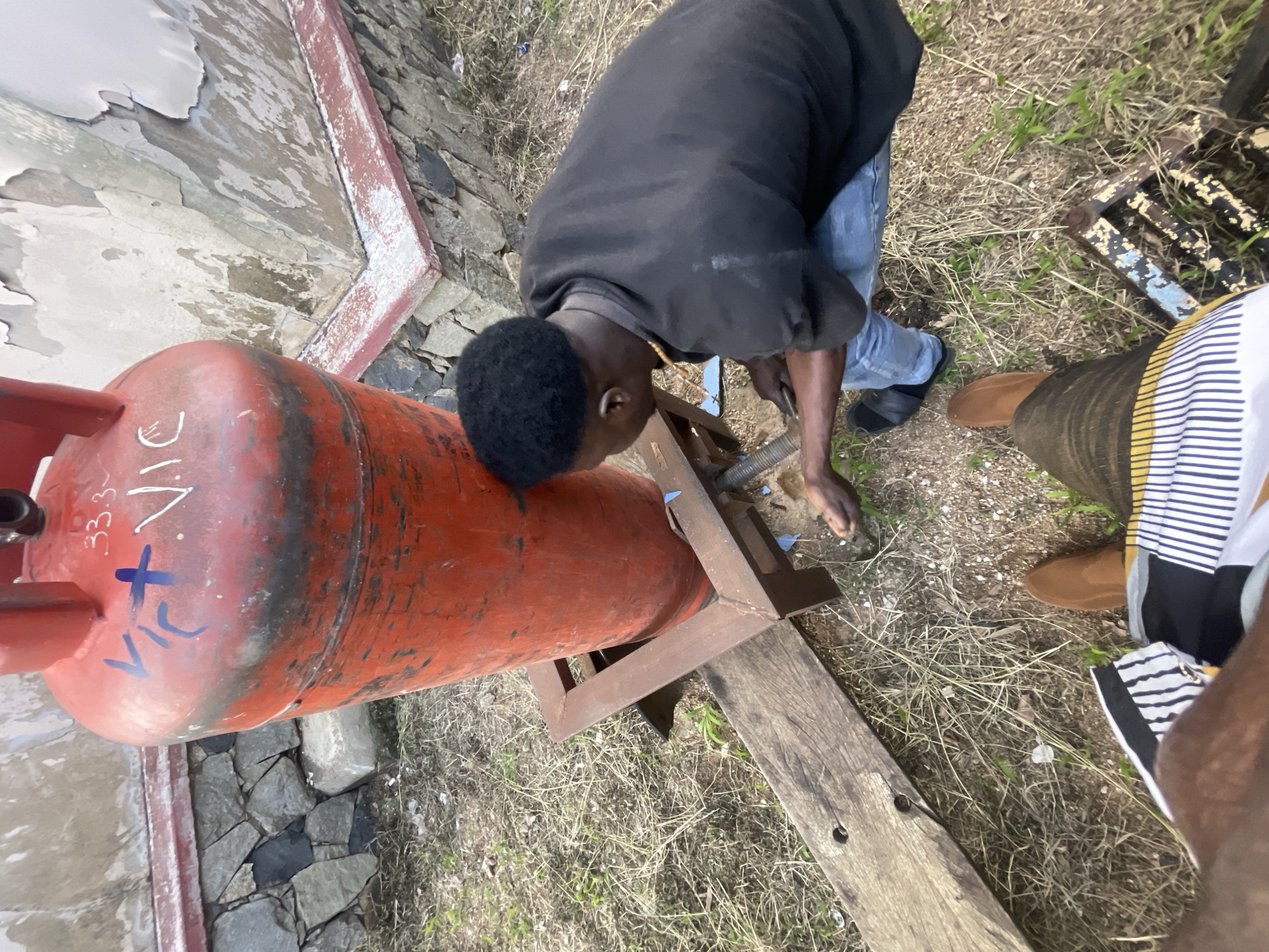 The image depicts a scene outdoors in Jos, Nigeria. The primary subject is a large, orange gas cylinder lying on its side, supported by a metal stand. A person, wearing a black long-sleeved shirt and blue jeans, is bent over the cylinder, seemingly working on it. Their head is angled downwards, obscuring their face. Another person, partially visible on the right, wears a striped shirt. The ground is covered with dry grass and loose stones. In the background, a rough, textured wall with peeling paint is visible, along with a stone foundation. The overall impression is one of a somewhat informal, possibly rural or semi-rural, outdoor setting. The lighting suggests it could be daytime, possibly late afternoon given the angle of the light, but the weather is not definitively discernible. Visible text on the gas cylinder, written in blue marker, includes "VIC+X" and "33.3~~". The activity appears to be related to the maintenance or use of the gas cylinder.