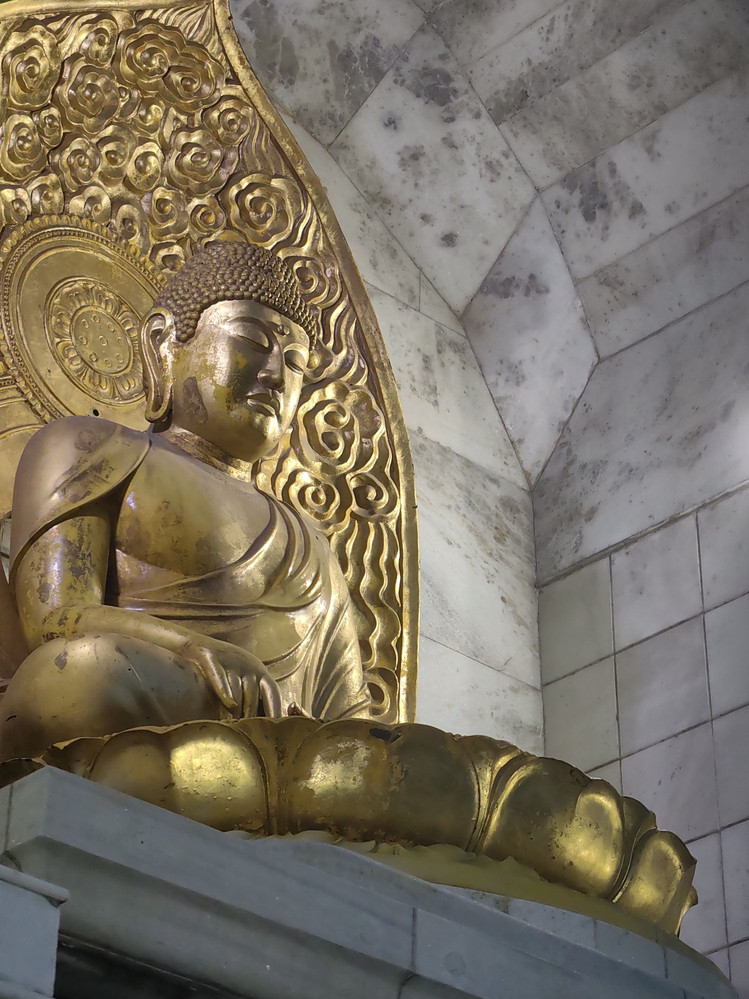 A golden statue of the Buddha is seated on a lotus pedestal, illuminated by warm light. The statue is positioned within an alcove with marble walls, the tiles exhibiting subtle variations in color and texture. Behind the Buddha's head is an ornate golden backdrop, intricately carved with floral and circular motifs. The statue's serene expression and the surrounding architectural elements suggest a place of worship or a cultural heritage site. This scene is likely located at the Lotus Temple in Delhi, India, known for its distinctive flower-shaped architecture and serene ambiance.
