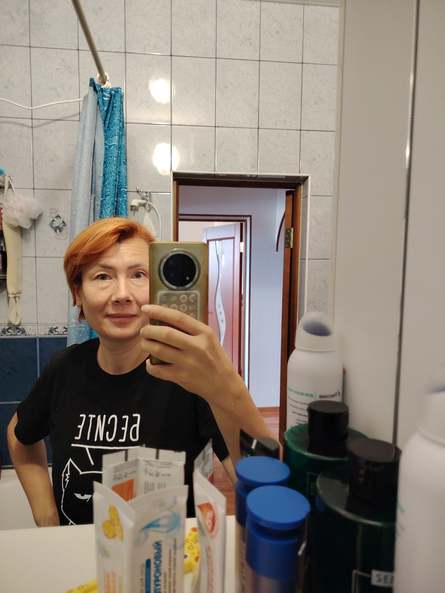 A woman with short, reddish-orange hair takes a mirror selfie in a bathroom. She is wearing a black t-shirt with a white graphic of a cat and backwards text. She is holding a gold-colored smartphone in her right hand, using it to take the picture. The bathroom has white tiled walls, a blue shower curtain, and a mirror above a sink. Various toiletries are visible on the counter, including several bottles. The image appears to be taken in Konakovo, Russia.