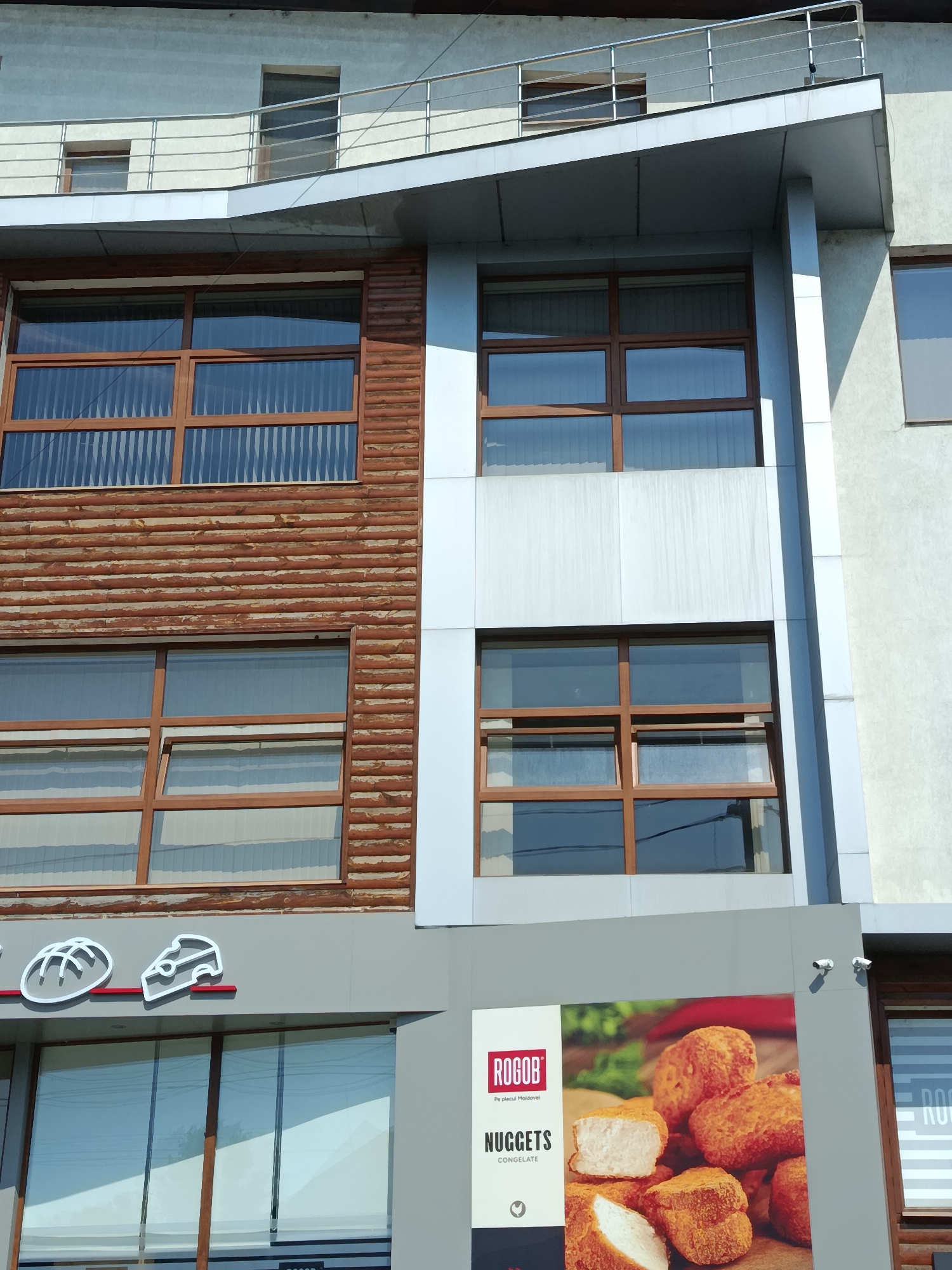 The image shows the exterior of a building in Chișinău, Moldova, with a focus on the facade. The building has multiple levels with distinct architectural features. The upper floors feature a grey, textured facade with horizontal bands and a metal railing. Below this, a section of the building is clad in horizontal wooden planks, which gives it a rustic appearance. This section contains several large windows with wooden frames, each divided into multiple panes. Adjacent to the wooden section is a part of the building with a lighter, possibly metallic or concrete, facade, also with windows.
In the lower part of the image, there is a grey signboard with graphics and text. The signboard displays a logo with stylized bread and cheese icons, and further to the right, a larger advertisement for "ROGOB NUGGETS CONGELATE" featuring an image of chicken nuggets. The building appears to be a commercial establishment, possibly a bakery or a fast-food restaurant, given the signage. Two small security cameras are visible mounted on the grey facade above the advertisement. The overall impression is of a modern building with a blend of contemporary and rustic design elements, located in an urban setting.