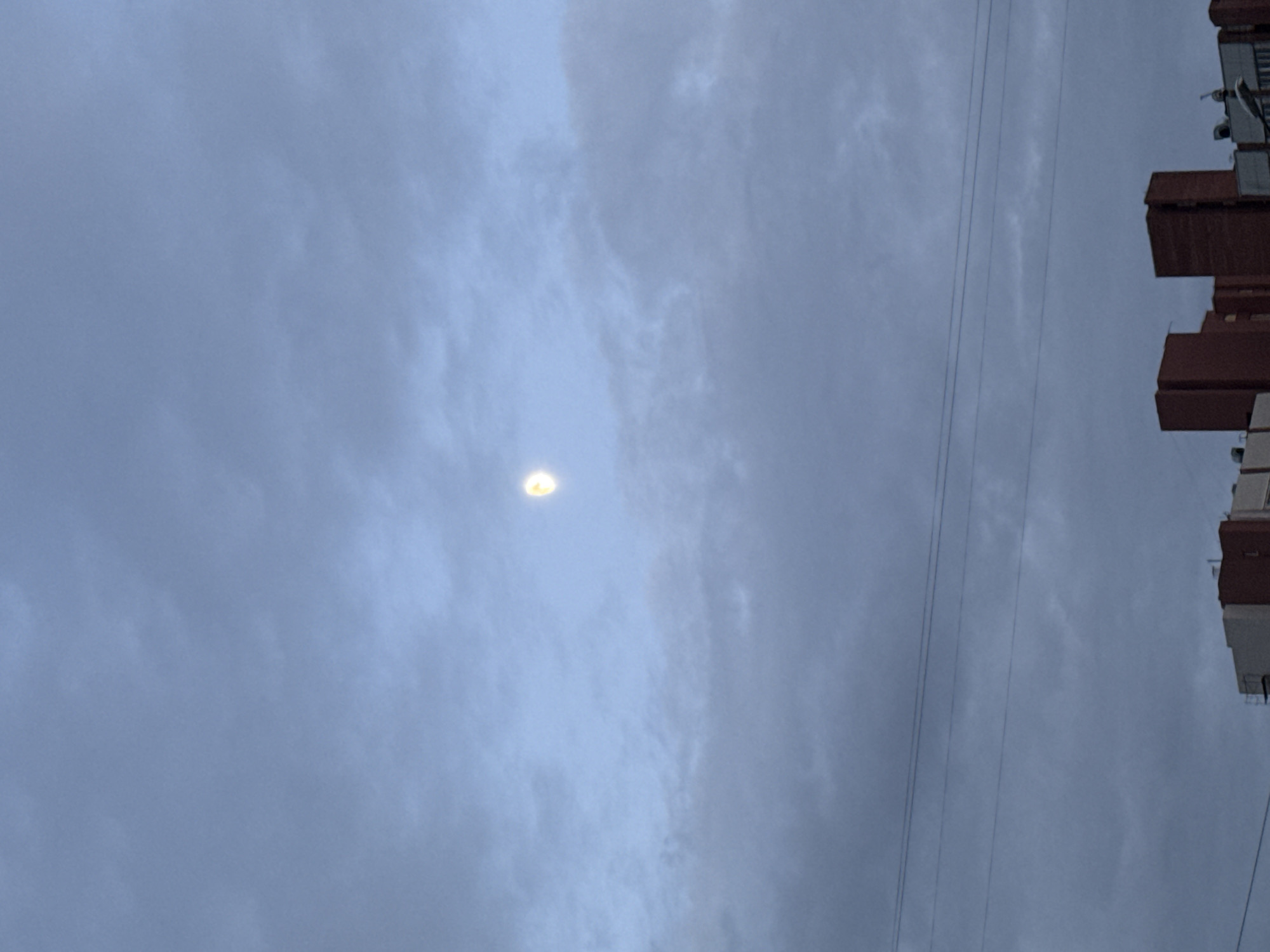 The image shows a sky with overcast clouds and a bright, round light source shining through the clouds. Power lines are visible on the right side of the image, along with the corner of a building. It is impossible to determine the location in the city of Saint Petersburg, Russia with the given information.
