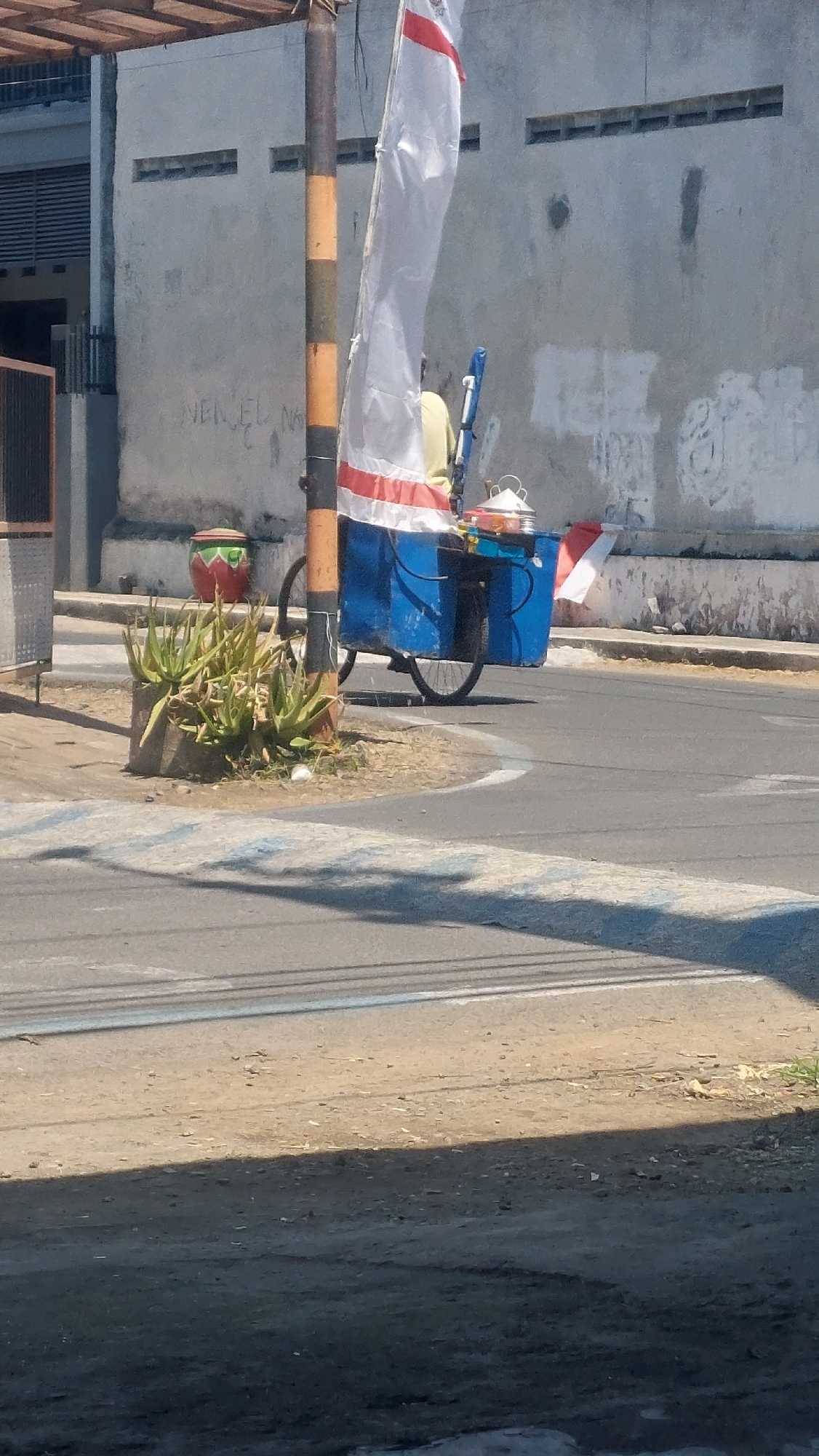 A tricycle food cart, painted blue and white with red accents, is parked on the side of a street in Sidoarjo, Indonesia. The cart is adorned with a tall, white flag that has red stripes. A person, wearing a yellow shirt, is seated on the cart, facing away from the camera. The cart's wheels are visible, suggesting it is mobile. In the foreground, a cluster of aloe vera plants grows in a concrete planter. Behind the cart, a weathered concrete wall with horizontal ventilation slits forms the backdrop. The ground is paved with asphalt, and a pedestrian crossing with blue and white stripes is visible. Sunlight casts shadows across the scene, indicating it is daytime.