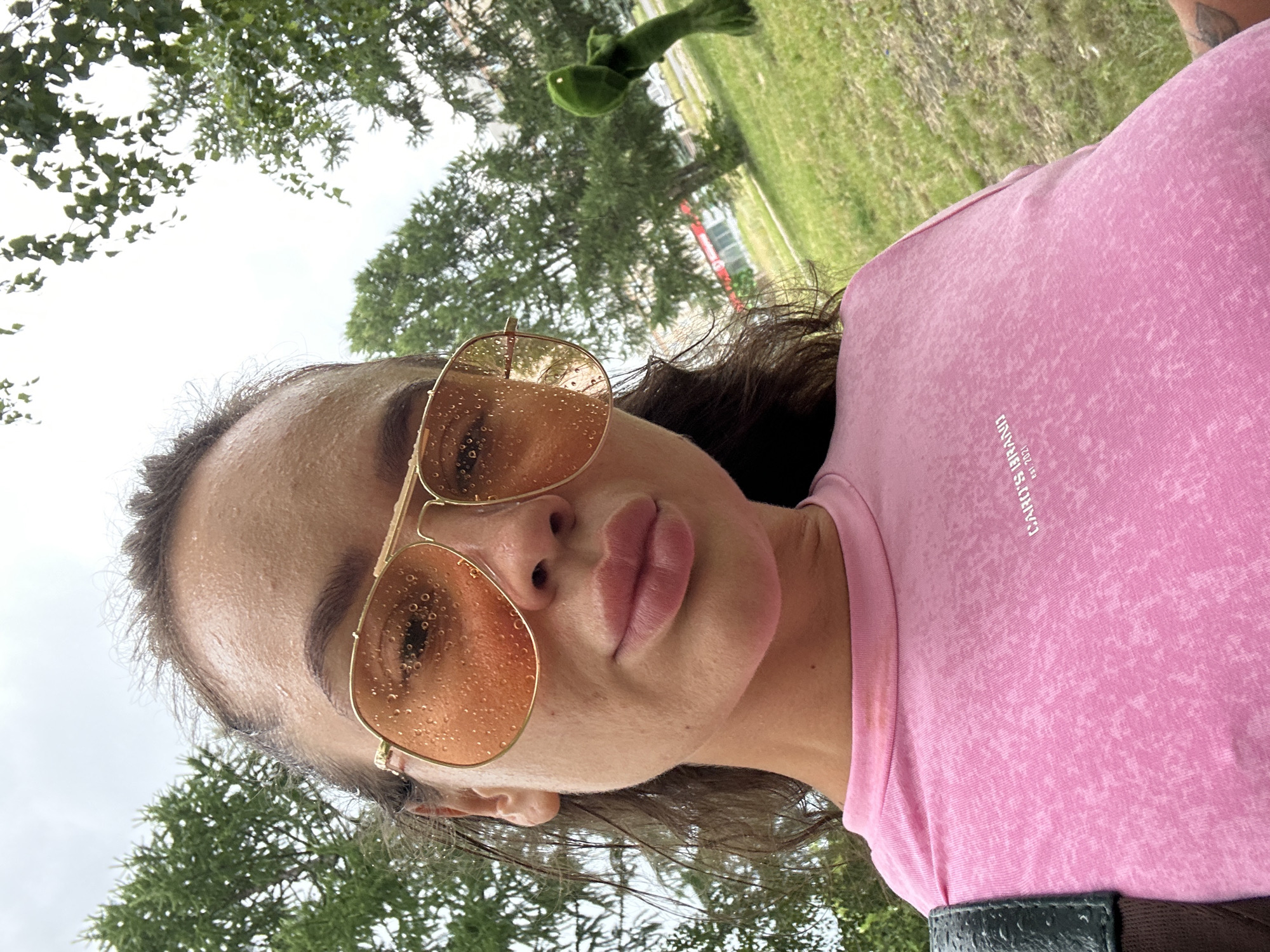 The image shows a close-up, low-angle shot of a person from the chest up, outdoors. The person is wearing a pink t-shirt with a textured, faded pattern and some faint white text that reads "CARO'S BRAND EST. 2021". They are also wearing large, aviator-style sunglasses with rose-tinted lenses, which are speckled with water droplets. The person's face is slightly glistening, suggesting it might be warm or humid, or perhaps they are sweating. Their lips are full and painted a soft pink. The background is blurred but shows greenery from trees and grass, with a cloudy sky above. In the upper left portion of the image, a blurry green toy frog is visible hanging from a tree branch. The scene appears to be in a park or grassy area. The lighting suggests it is daytime, likely overcast or after rain due to the water droplets on the sunglasses and the glistening skin. The overall mood is casual and relaxed. The image was taken in Yuzhnouralsk, Russia.