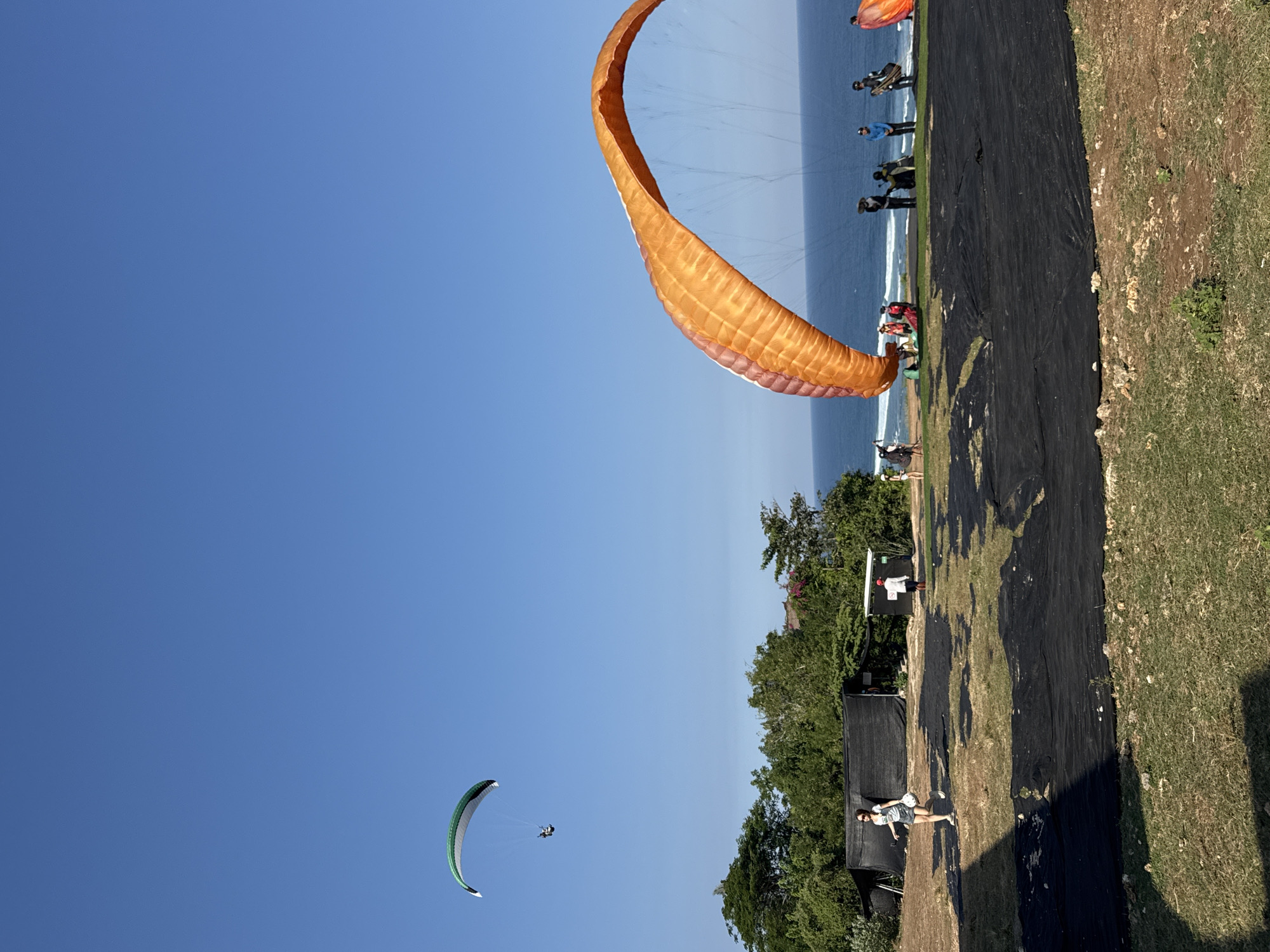 In Kuta Selatan, Indonesia, on a sunny day, paragliders prepare for or are in the process of flight. A paraglider with a green and white canopy is soaring high in the clear blue sky. In the foreground, a large orange and white paraglider wing is spread out on the ground, with several people attending to it and potentially preparing for their own flights. Other individuals are scattered around the grassy takeoff area overlooking the ocean. The landscape features lush green trees and vegetation leading down to the blue sea. Black tarps are laid out on the ground, likely to protect the grass or for the paragliders to use. The scene captures the activity and excitement of paragliding in a scenic coastal location.