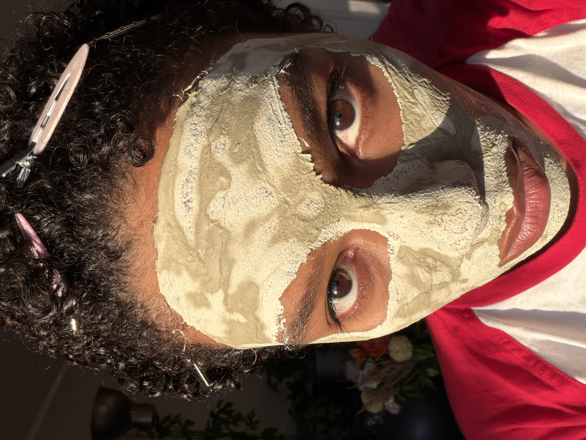 The image is a close-up, high-angle shot of a person wearing a clay face mask. The person has curly dark hair with several hair clips, some metallic and some pink, holding sections of hair back. The face mask is light beige and covers most of the face, revealing only the eyes, nose, and lips. The person's eyes are brown and looking directly at the camera. The lips are painted with a reddish-brown lipstick. They are wearing a red and white baseball-style t-shirt. In the background, out of focus, there is some foliage and a metallic object that could be a lamp. The lighting suggests natural sunlight coming from the right, casting shadows on the left side of the face. There is no indication of the city of Champs-sur-Marne, France, in this image.