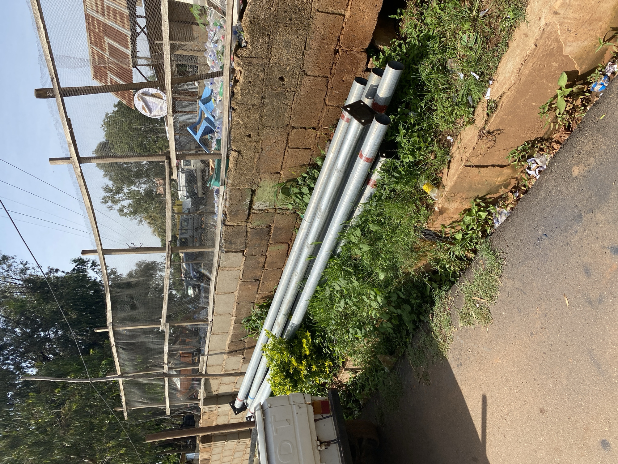 The image depicts an outdoor scene in Gura Top, Nigeria, featuring a pile of metal pipes lying on the ground next to a brick wall. A portion of a white truck is visible on the left side of the frame. Beyond the wall, a fence made of wooden posts and mesh encloses an area with trees and some structures, including a building with a corrugated metal roof. Scattered plastic bottles and debris are visible in the fenced area and along the roadside ditch. The ground is a mixture of asphalt and dirt, with grass and vegetation growing in patches. The sky is clear and blue.