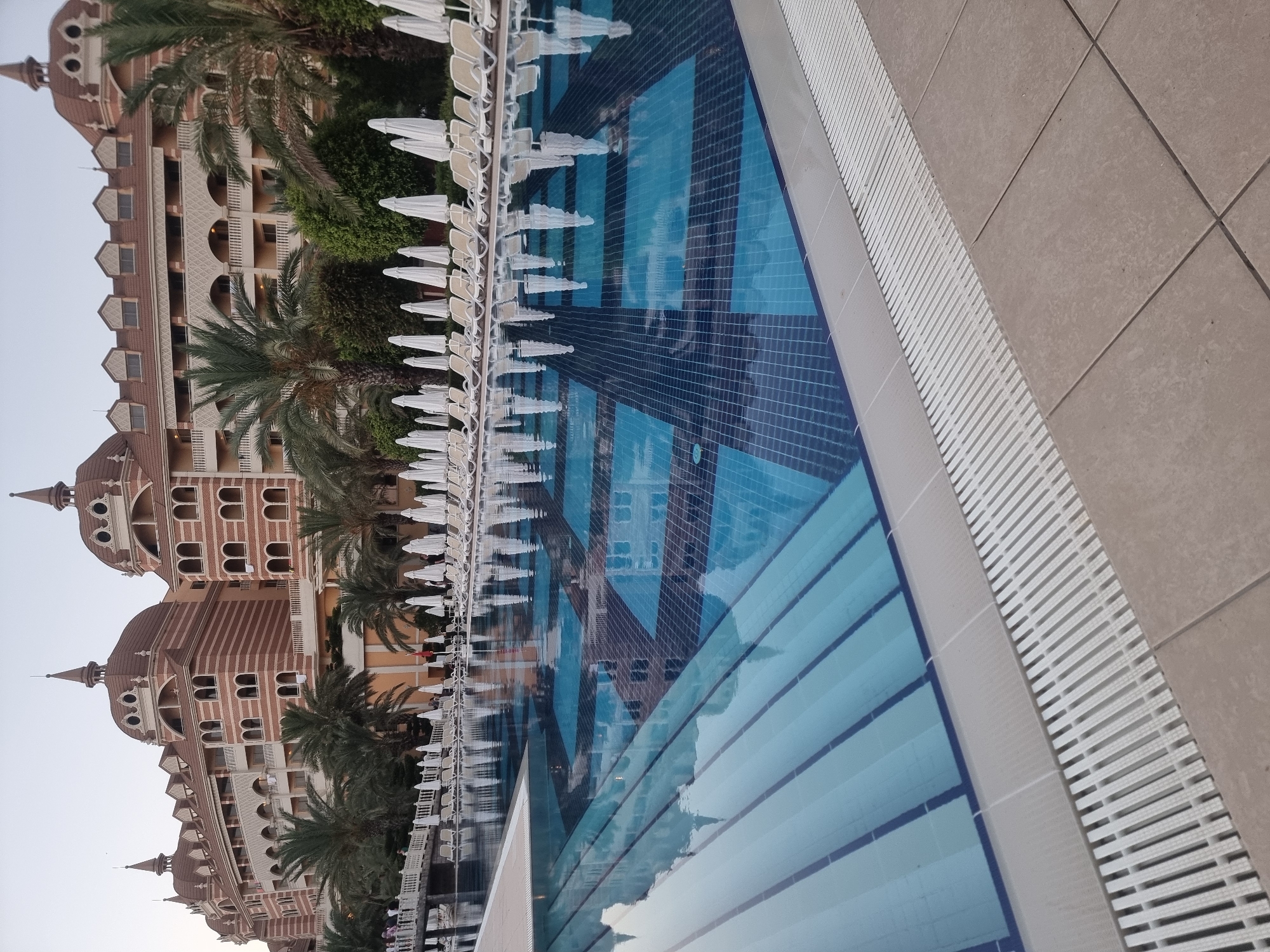 The image depicts a large swimming pool in front of a hotel in Manavgat, Turkey. The hotel is a multi-story building with ornate architecture, featuring arched windows and domed roofs, and is constructed with red and beige brick. Palm trees and lush green bushes surround the hotel and pool area. White lounge chairs and closed umbrellas are neatly arranged along the edge of the pool. The swimming pool itself has a blue tiled bottom with a patterned design. The water reflects the hotel, trees, and sky. The foreground shows the edge of the pool and the paved deck area, with a white grating system for drainage. No people are clearly visible engaging in any specific actions within the pool or on the deck, though a few figures are faintly visible on a walkway in the background, suggesting a resort setting. The sky is a pale, overcast hue, indicating it might be early morning or late evening.