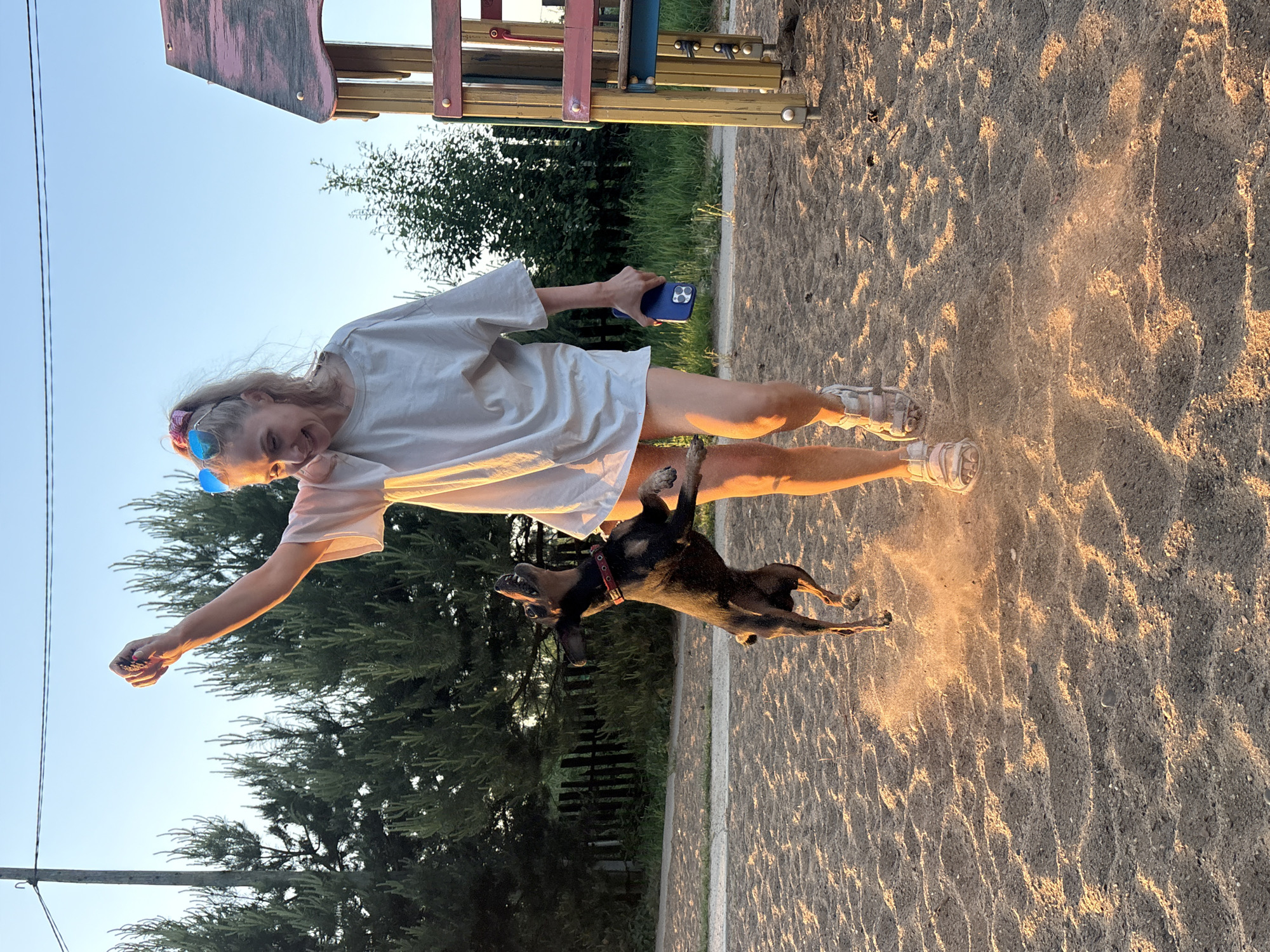 A fair-skinned woman with blonde hair is playing with a small black dog in a sandy playground, possibly in the city of Chaikovsky, Russia. The woman is wearing a white oversized t-shirt, sandals, and sunglasses pushed up on her head. She is holding a blue phone in one hand and extending the other hand towards the dog, which is leaping up into the air. The playground appears to be surrounded by trees. The photograph is taken at dusk, casting long shadows across the sand.