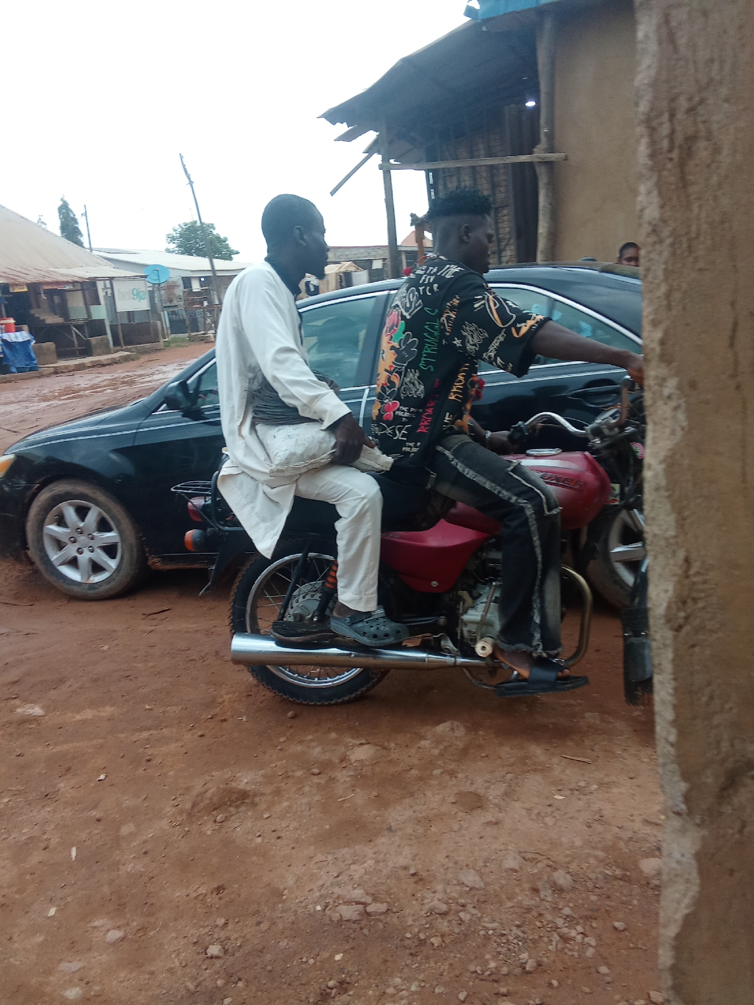 Two men ride on a red motorcycle. The man in front, wearing a white garment, sits with his back to the viewer. The man behind, wearing a patterned black shirt and jeans, drives the motorcycle. They are on a dirt road next to a black car and buildings. The sky is overcast.
