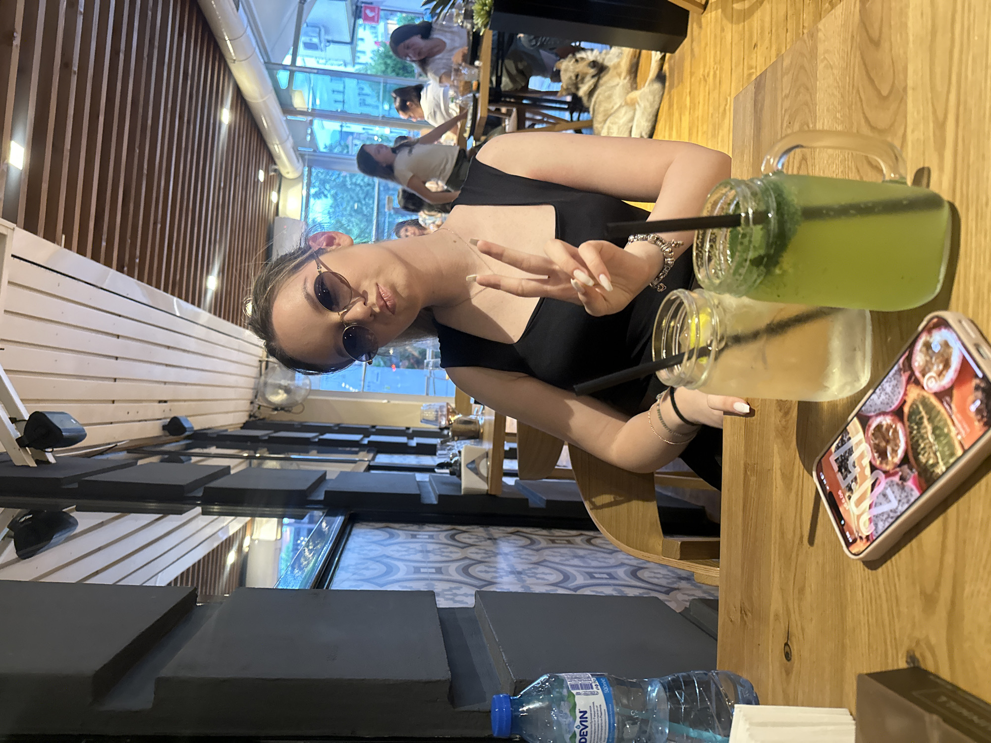 The image captures a young woman seated at a wooden table inside **Porto Maltese Restaurant in Blagoevgrad, Bulgaria**, during the early evening, as indicated by the "20:31" time displayed on a smartphone.
The main subject, a young woman with light brown hair pulled back, wears dark, round sunglasses and a black square-neck top. She has a subtle "duck face" expression and holds her right hand up, forming a peace sign (V-sign) with her manicured, light-colored nails. She wears a delicate necklace and a charm bracelet on her left wrist, along with multiple thin bracelets on her right wrist.
On the light-colored wooden table in the foreground are two drinks in large mason jar mugs with black straws: one clear, lemon-infused drink and one vibrant green, mint-infused drink. To her left, a half-full 500ml bottle of "DEVIN" branded mineral water with a blue cap is present. A white napkin dispenser and a dark brown rectangular object (possibly a wallet or small box) are also on the table. A light-pink cased smartphone lies face-up, displaying its lock screen with the time "20:31" and a colorful wallpaper featuring sliced exotic fruits like passion fruit, dragon fruit, and kiwi.
The restaurant's interior features a distinctive design. To the left, a wall is adorned with grey and white/light blue patterned tiles, framed by dark grey architectural elements. The ceiling is characterized by light wooden slats interspersed with dark beams, from which black speakers are suspended. In the mid-background, other patrons are visible seated at wooden tables, engaged in conversation. A large, light-colored, wolf-like dog lies calmly on the light wooden floor to the right of the main subject, near another table. Through large windows in the background, an urban environment is visible, with other buildings and a glimpse of a street scene, consistent with a city location.