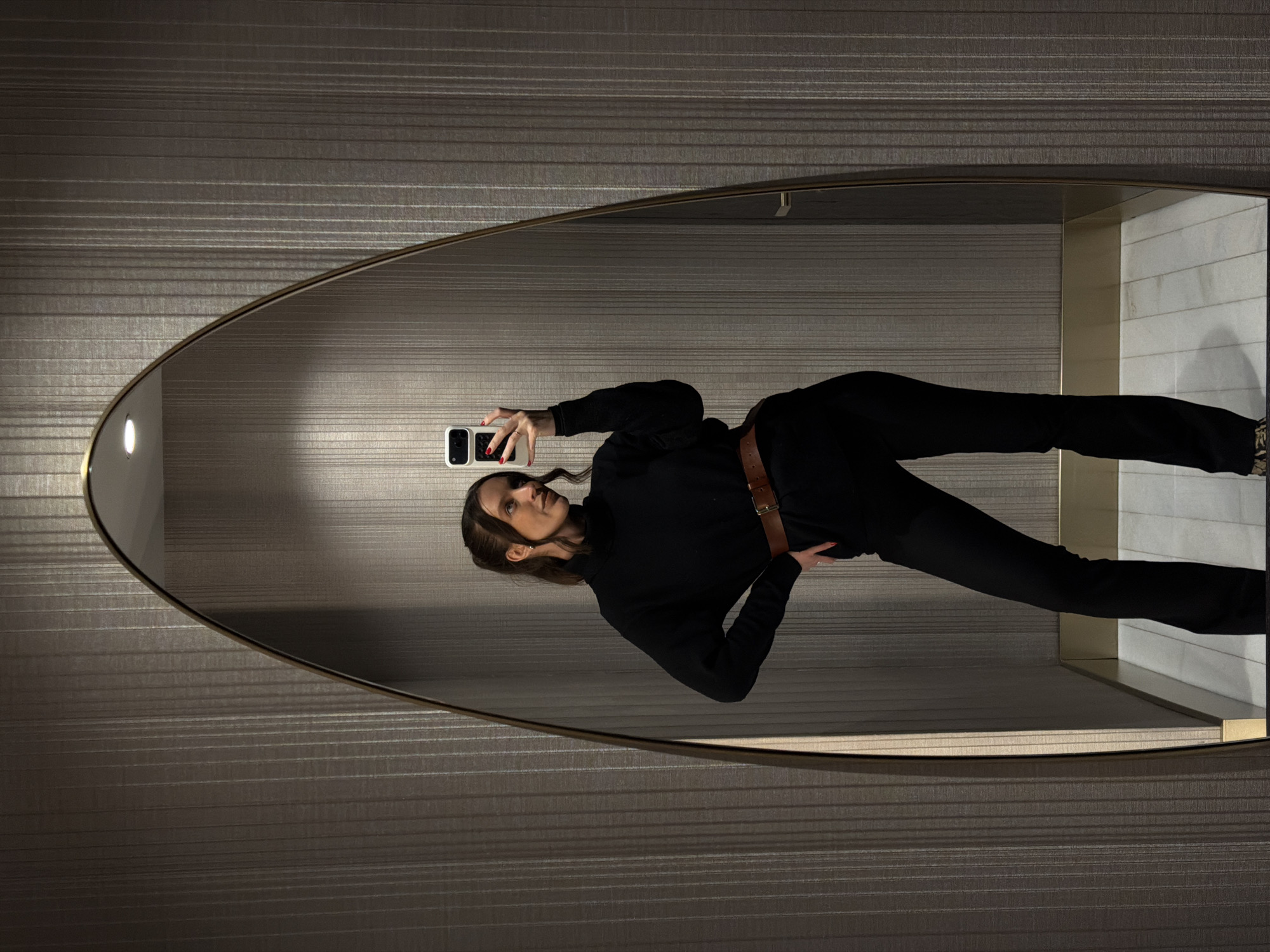The image depicts a female individual captured in a mirror selfie within an interior space in Budva, Montenegro, likely a modern dressing room or hotel room. The subject, a woman with dark hair pulled back into a half-up style, wears a black turtleneck sweater, black trousers, and a brown leather belt with a gold-toned buckle. Her left hand is placed on her hip, while her right hand holds a white smartphone with a dark multi-lens camera module, pointed towards the mirror to capture the reflection. Her fingernails are painted red. She wears small, clear earrings. The mirror is tall and irregularly shaped with an arched top, framed by a thin gold-colored border. The surrounding walls are covered in dark grey textured wallpaper featuring fine vertical striations. The floor visible in the reflection is light-colored, possibly white or light grey, with a tiled pattern. A gold-toned trim separates the wall from the floor. A recessed light fixture is visible in the ceiling reflection within the mirror's upper area, and a dark door handle is faintly discernible in the mirror's right side.
