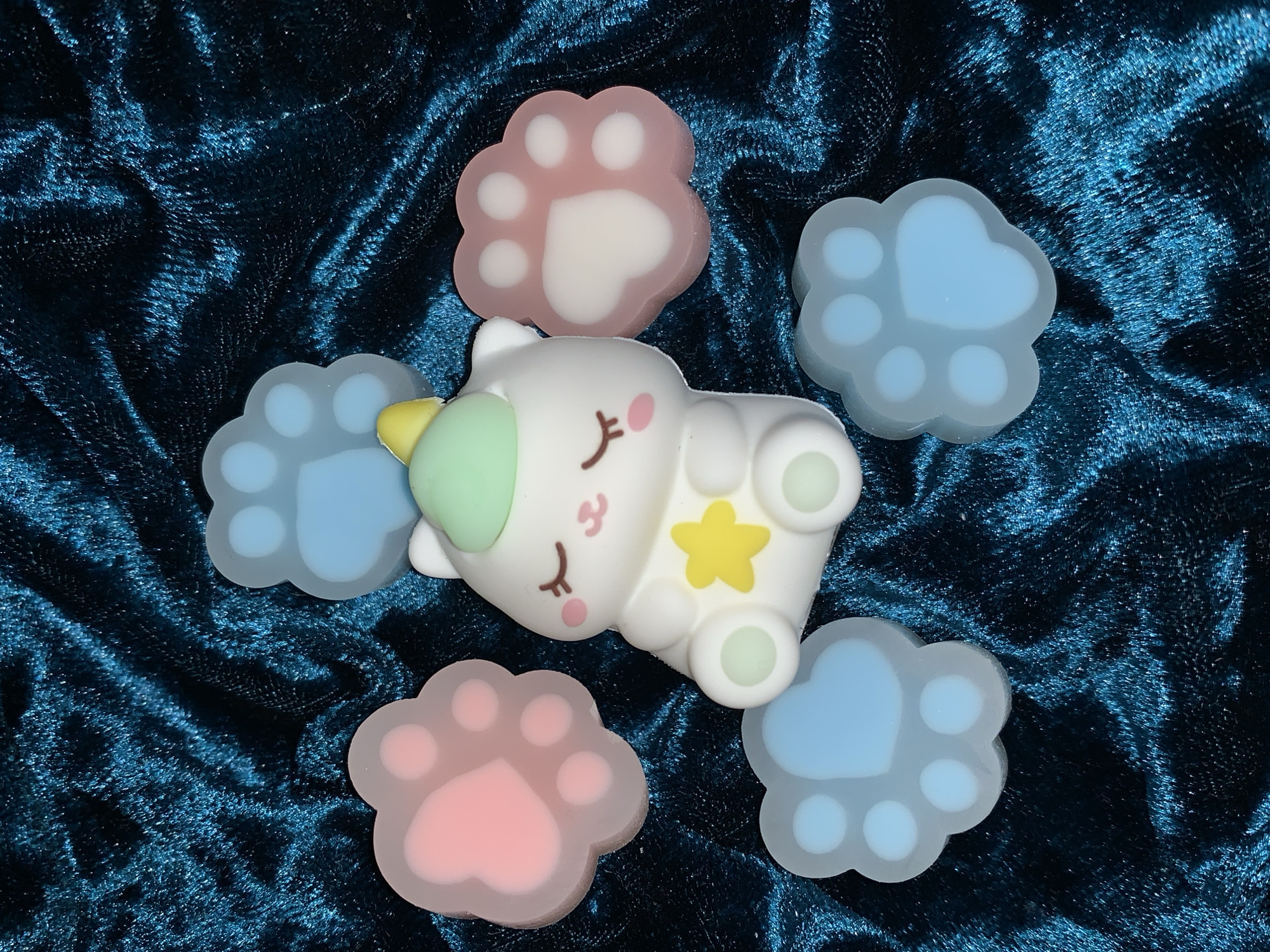 The image displays a whimsical arrangement of small, toy-like objects on a dark teal or blue-green crushed velvet fabric. The fabric forms soft folds and ripples, creating a textured background with varying shades due to light reflection.
The central subject is a white, cartoon-style character resembling a sleeping unicorn-cat hybrid, often called a "caticorn." It lies on its back with its eyes closed, featuring delicate eyelashes and rosy pink cheeks. A small, subtle smile is visible on its face. It has a light green horn with a yellow tip and a yellow star emblem on its body. Its four paws, visible at the ends of its limbs, appear to have light green circular pads.
Arranged around the caticorn are five paw-print shaped items. Three of these paw prints are a soft, translucent light blue, each featuring a larger heart-shaped central pad and four smaller circular toe pads. The remaining two paw prints are a similar soft, translucent light pink, also designed with a heart-shaped central pad and four circular toe pads.
The overall scene suggests a gentle and playful aesthetic, emphasizing cuteness and soft colors against the rich, deep hue of the velvet. There is no discernible activity taking place, and no people or text are visible in the image. The setting appears to be indoors, showcasing these decorative or collectible items.