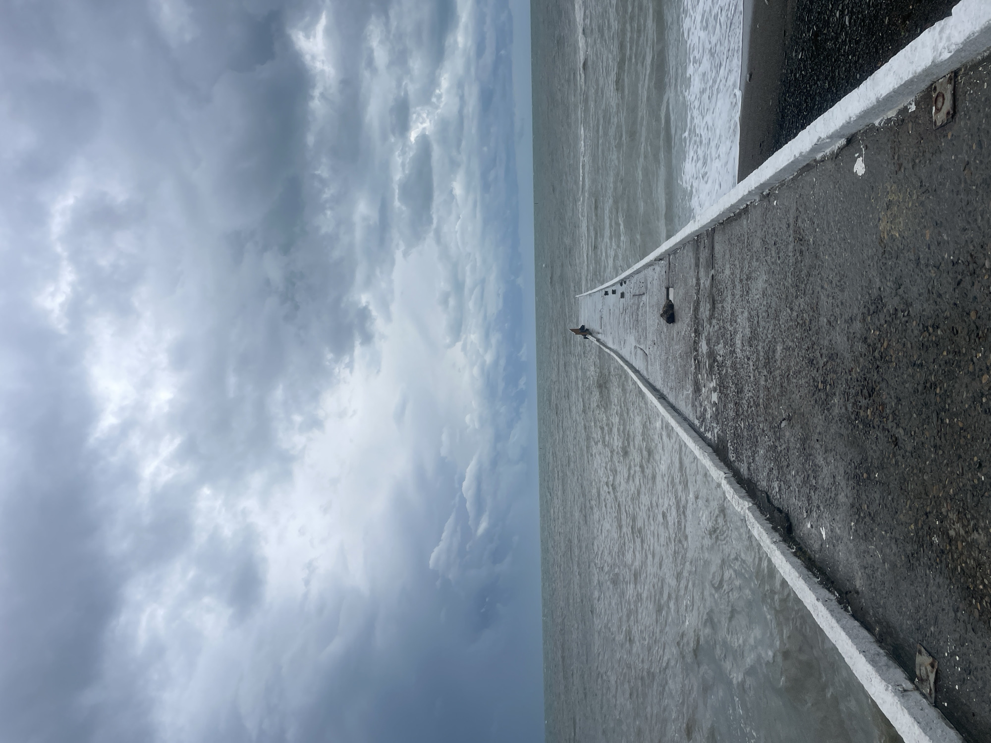 The image captures a pier extending into the sea, presumably in Sochi, Russia, though that is not discernable from the image alone. The pier is made of concrete, with a white-painted curb along the edges. The sea is choppy, with waves visible near the shoreline. The sky is overcast with dark clouds, suggesting an impending storm. There are a few dark objects on the pier that may be birds. The perspective is from a low angle, looking out towards the horizon. The image is oriented sideways, with the sky on the left and the pier on the right.