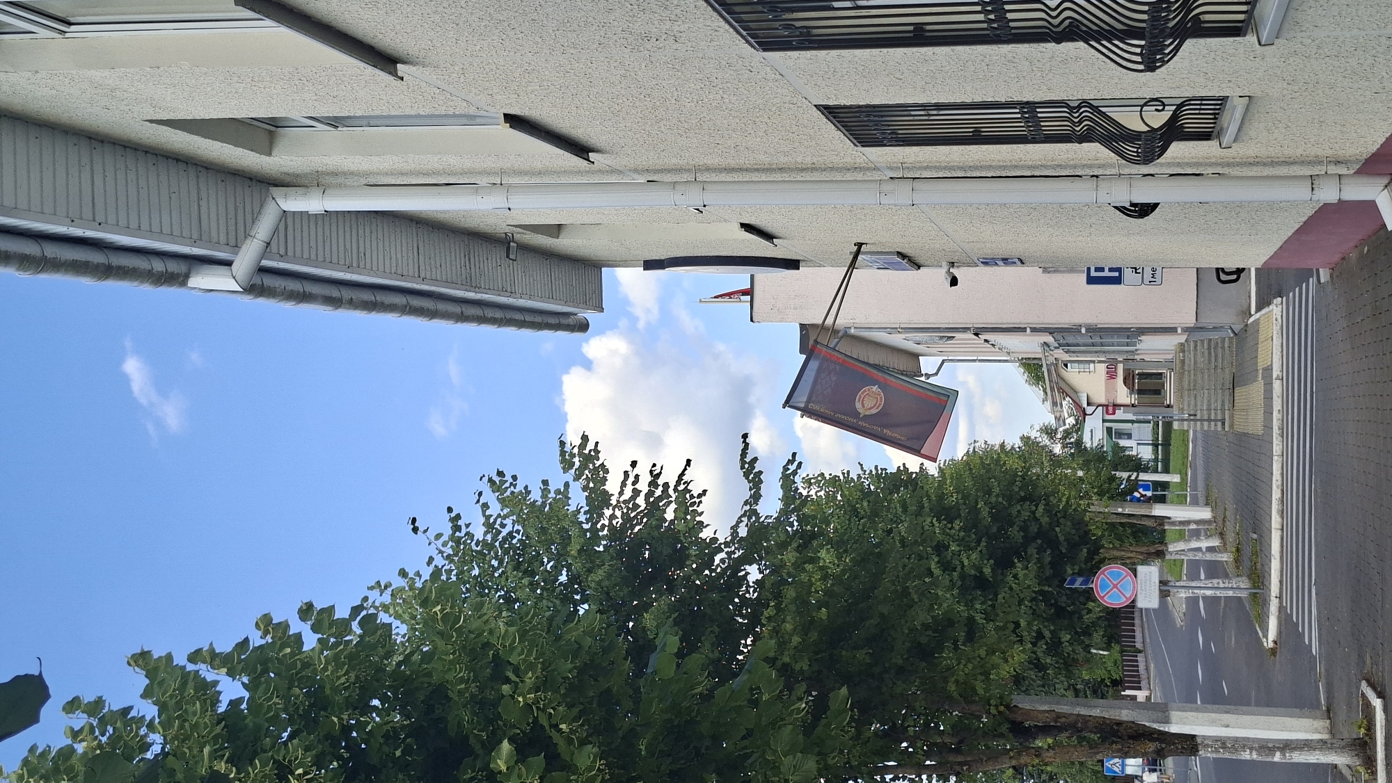 The image shows a street scene in the city of Myadzyel, Belarus, on a partly cloudy day. In the foreground, a sidewalk runs along the side of a light-colored building. A flag, featuring a dark red and black pattern with a circular emblem in the center, is displayed on a pole extending from the building. The flag has Cyrillic writing on it. A white drainpipe runs vertically along the building's exterior. To the left of the building, a line of trees with dense green foliage is visible, their trunks painted with white at the base. A street with a crosswalk is also present, with traffic signs and a pedestrian crossing symbol visible. In the distance, other buildings and more trees can be seen. The sky is blue with scattered white clouds. No people are clearly visible in the image.