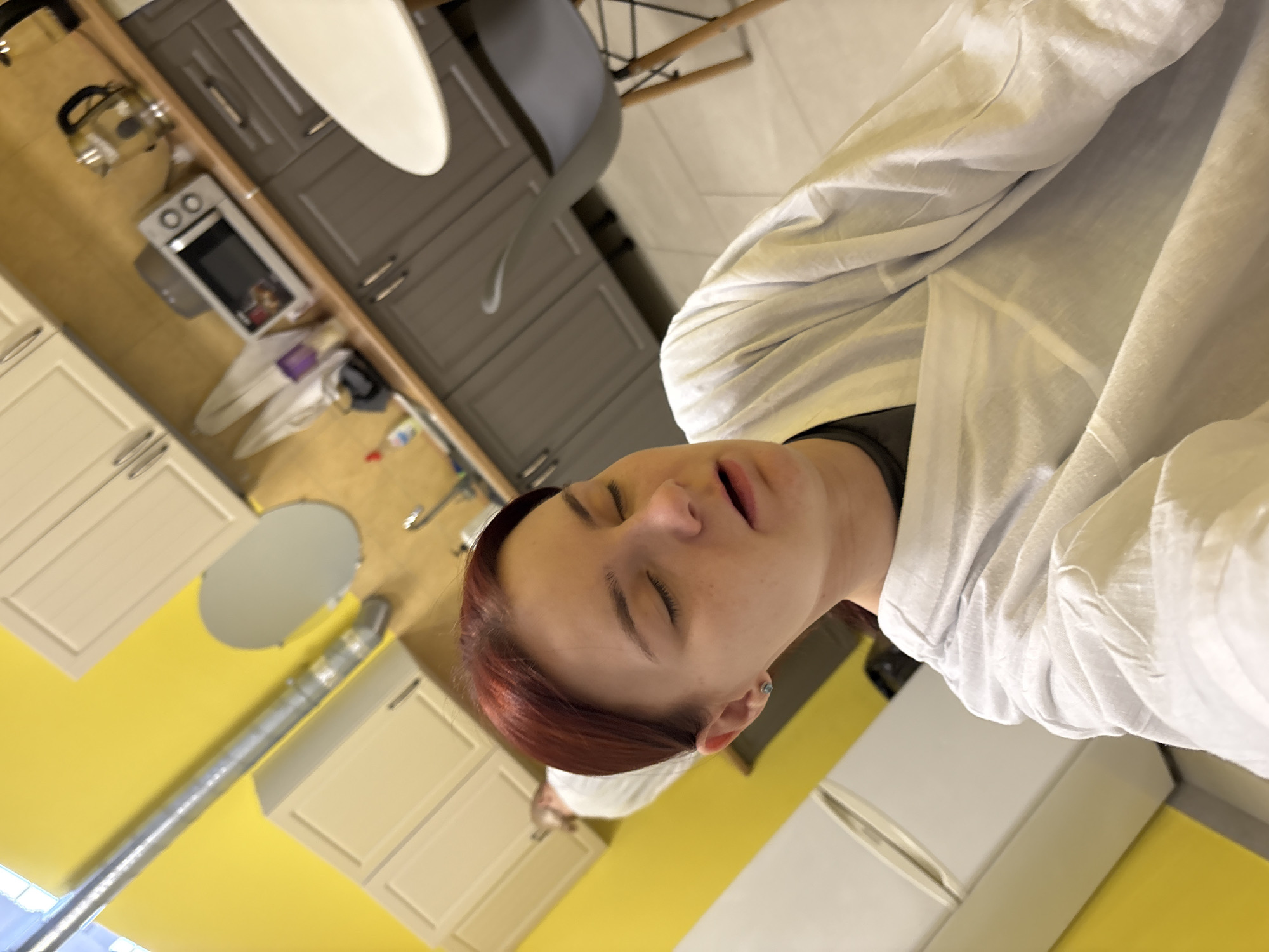 A medium shot captures a young Caucasian female with auburn hair and closed eyes, her mouth slightly open, positioned in the foreground. She is wearing a white, unbuttoned lab coat over a dark gray shirt. The background reveals a brightly lit kitchen area with yellow walls. A metal vent pipe runs diagonally across the upper left of the frame. The kitchen features light-colored upper cabinets and darker gray lower cabinets. On the countertop, a microwave, a kettle, and various other items are visible. To the right, a white tabletop and a gray chair with wooden legs are present. Further back, another person, also wearing a white garment, stands near the cabinets, their face obscured. The scene suggests an indoor setting, possibly a laboratory or a communal kitchen space.