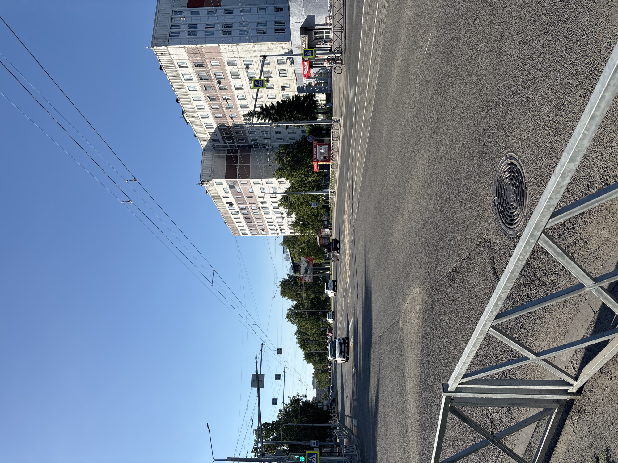 The image depicts a street scene in Novokuznetsk, Russia, under a clear blue sky. A wide asphalt road with multiple lanes stretches into the distance, bordered by sidewalks and buildings. Cars are visible driving on the road, and a pedestrian crossing with a green traffic light is present on the left side of the image. Overhead, power lines crisscross the sky, supported by poles. On the right side of the road, a large, multi-story residential building with many windows dominates the background. In front of the building, there are a few small kiosks and bus stops. A metal fence runs along the edge of the sidewalk in the foreground. The scene appears to be a typical urban environment on a sunny day.