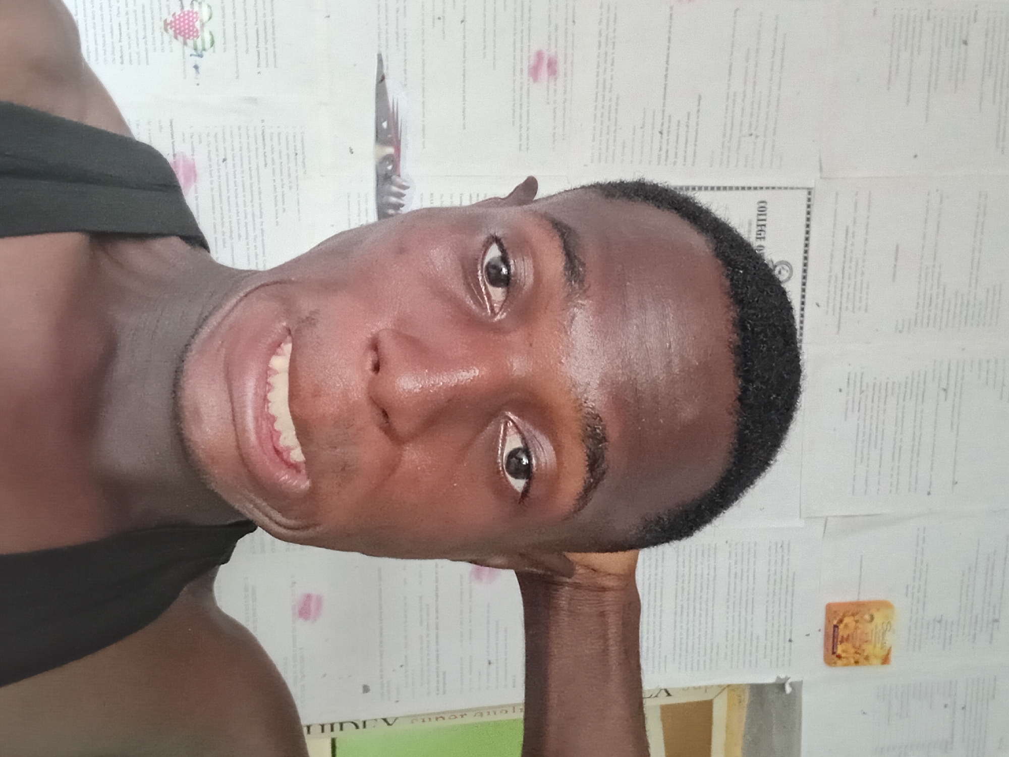 A medium-shot, close-up portrait of a dark-skinned man with short, dark hair. He is smiling and looking directly at the camera. He is wearing a black athletic strap. The background consists of sheets of paper with printed text and a small, tan-colored, rectangular object. The location is not identifiable as Akwete, Nigeria, based on the given visual information.