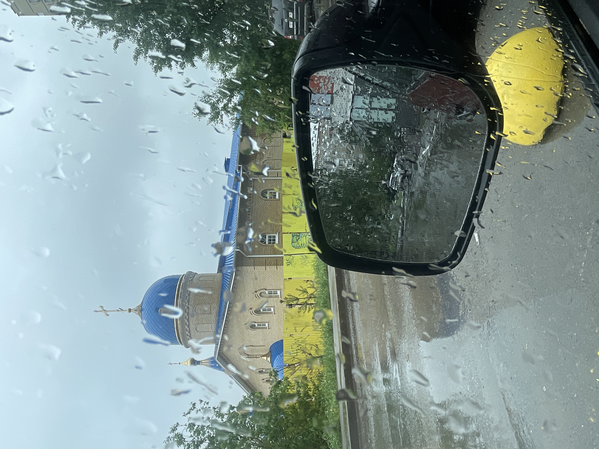 The image shows a rainy day scene through a car windshield, with water droplets obscuring the view. A side mirror is visible on the right side of the frame, reflecting the surroundings. On the left side, a building with a blue dome resembling a Russian Orthodox church is seen, possibly undergoing renovation or construction, as scaffolding or protective material is evident around it. The environment appears urban, with trees and other buildings in the background. The overall setting suggests a city, potentially in a region with Russian or Eastern European architectural influences.