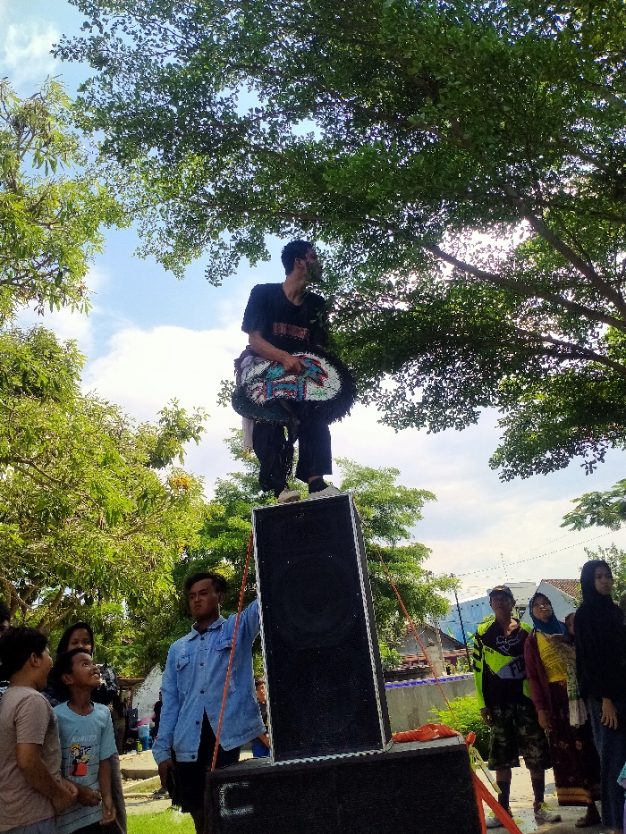 An outdoor scene in Jetak, Indonesia, depicts a male performer standing atop two stacked black speaker boxes. He wears a black t-shirt, dark pants, white shoes, and an ornate, multi-colored, circular adornment around his waist. His face and neck show traces of green material. He looks upwards and to the right, mouth open, suggesting a vocal performance. A young male in a light blue denim jacket stands near the speaker boxes with his hand on the upper one. To the left, two young males and a woman observe the performer. To the right, a man in a neon yellow/green and black shirt, a woman holding a baby, and another woman in a dark headscarf are among the onlookers. The setting is beneath a canopy of large green trees, with a bright blue sky and white clouds visible above. In the background, white buildings and a blue wall are present.