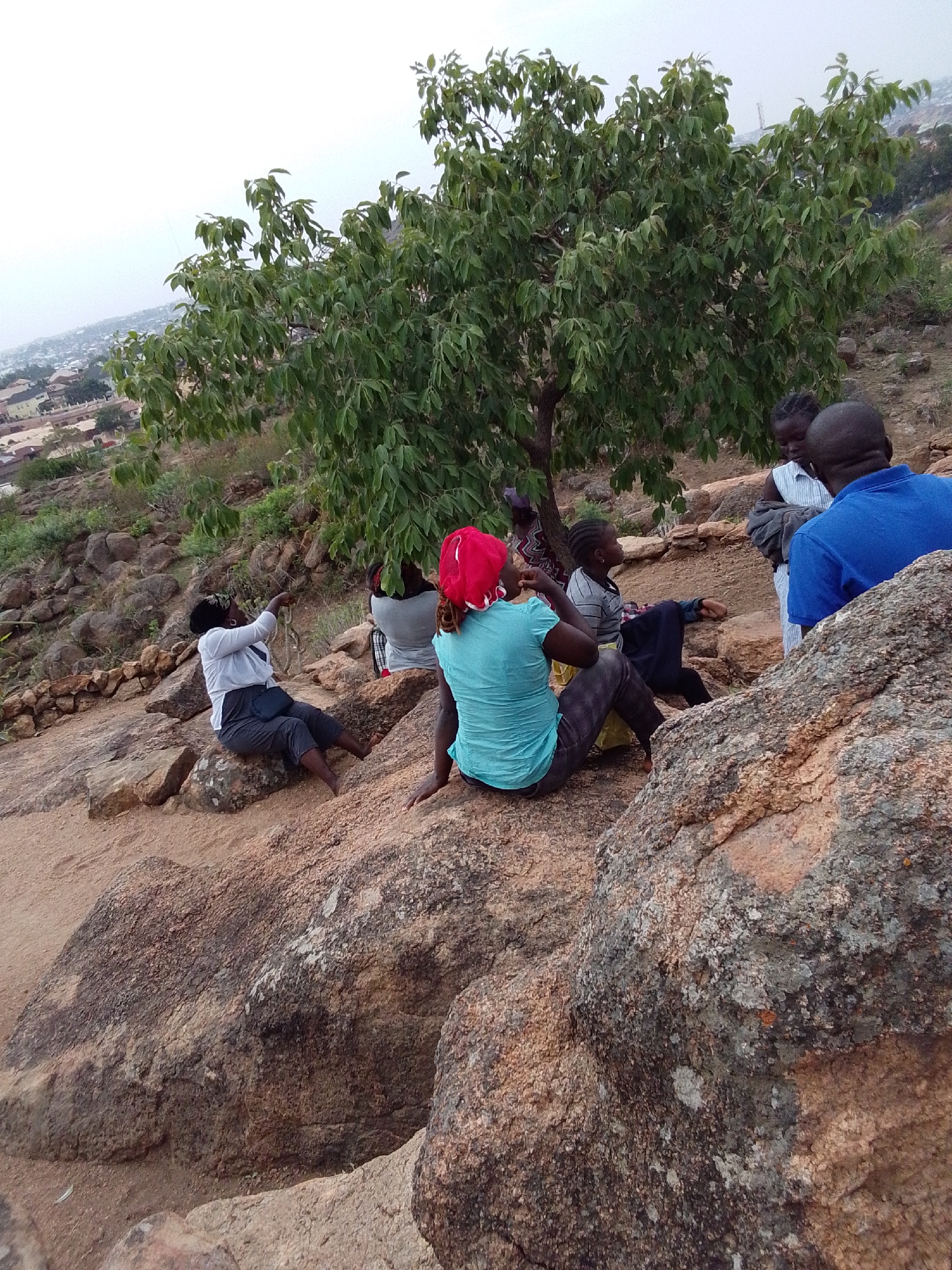 The image shows a group of people gathered on a rocky, sandy outcrop. A tree with lush green foliage grows nearby. The city of Gura Top, Nigeria, is visible in the background, with buildings and vegetation scattered across the landscape under a cloudy sky.
Several individuals are seated or standing among the rocks. One person is seated facing away from the camera, wearing a light blue shirt and a red head covering. Another individual is seated further to the left, wearing a light-colored shirt and dark pants. Two other individuals are standing, one wearing a blue shirt and the other a light-colored striped shirt. The group appears to be engaged in conversation or relaxation. The scene suggests a natural, outdoor environment.