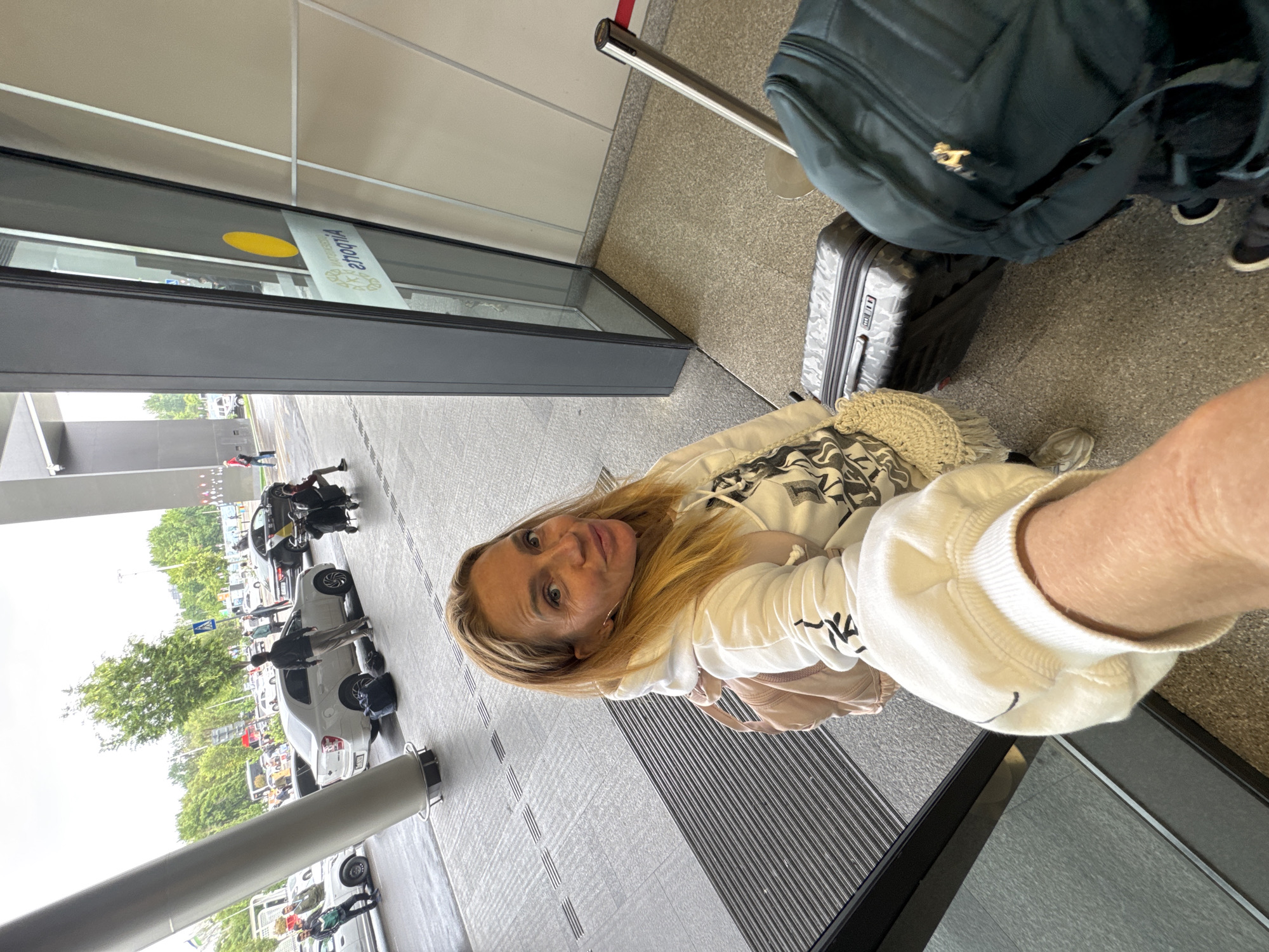 A medium indoor shot shows a woman in front of a glass building in Tashkent, Uzbekistan, with luggage beside her.
The woman is fair-skinned with blonde hair. She wears a white sweatshirt with black graphic designs on the sleeves. Her face is visible, and she is looking at the camera.
Beside her is a gray suitcase and a green backpack. A stanchion post with a red stripe is also visible. The building appears to be an airport or similar transportation hub, with a sign visible on the glass.
Outside the building, cars, trees, and people can be seen.