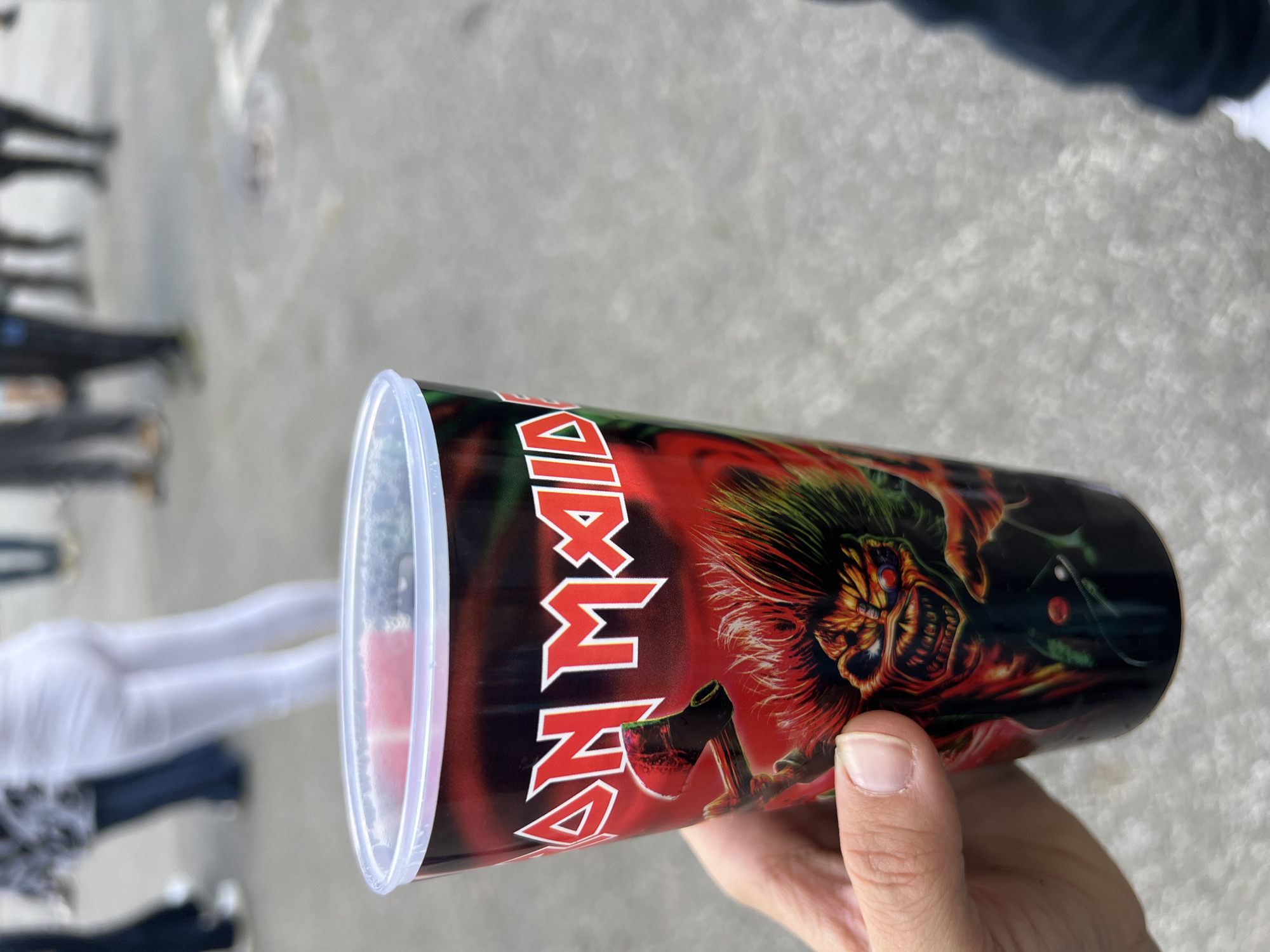 A close-up, outdoor shot shows a hand holding a plastic cup with an image of Iron Maiden's "Eddie" on it. The cup is filled with a red liquid. In the blurred background, there are legs of people walking. The scene appears to be in a public space, potentially in Sofia, Bulgaria, though there is no visual confirmation of it.