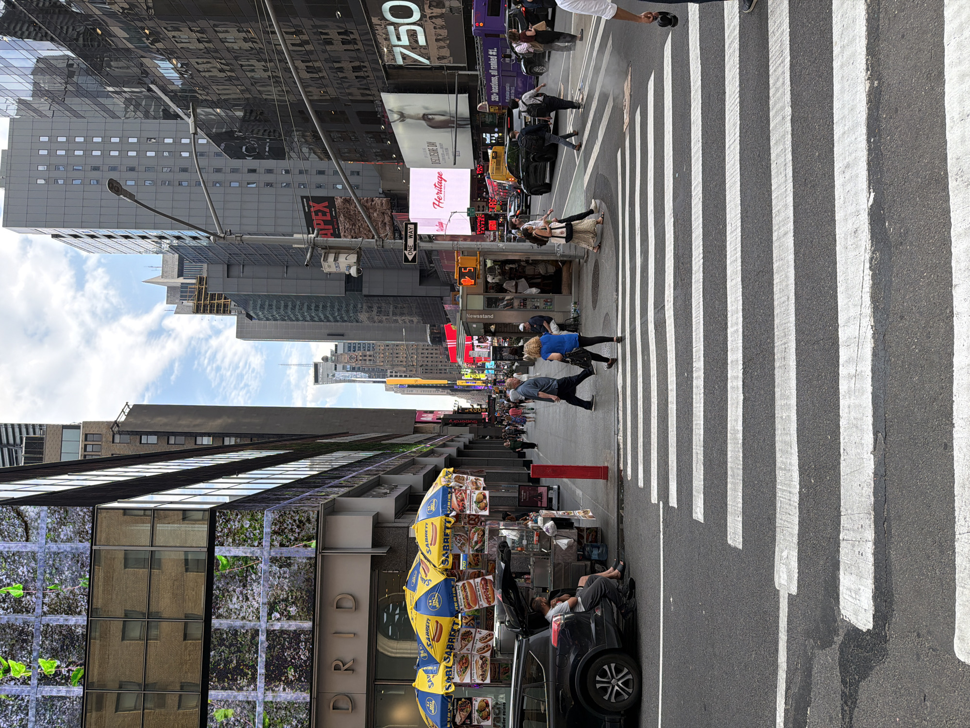 This is a street-level view looking down a New York City street, United States of America. The image showcases elements of city life, including pedestrians crossing in a crosswalk and street vendors.
A hot dog cart adorned with "Sabrett" umbrellas stands on the sidewalk. A car is parked nearby, with someone inside. Pedestrians are actively crossing the street. Buildings flank both sides of the street, with signs and advertisements visible. Some of the signs read "750," "Heritage DISCLOSURE DAY," "APEX," and "ONE WAY." The facade of a building features a design with foliage. A newsstand is present on the sidewalk. The sky is partially visible between buildings.