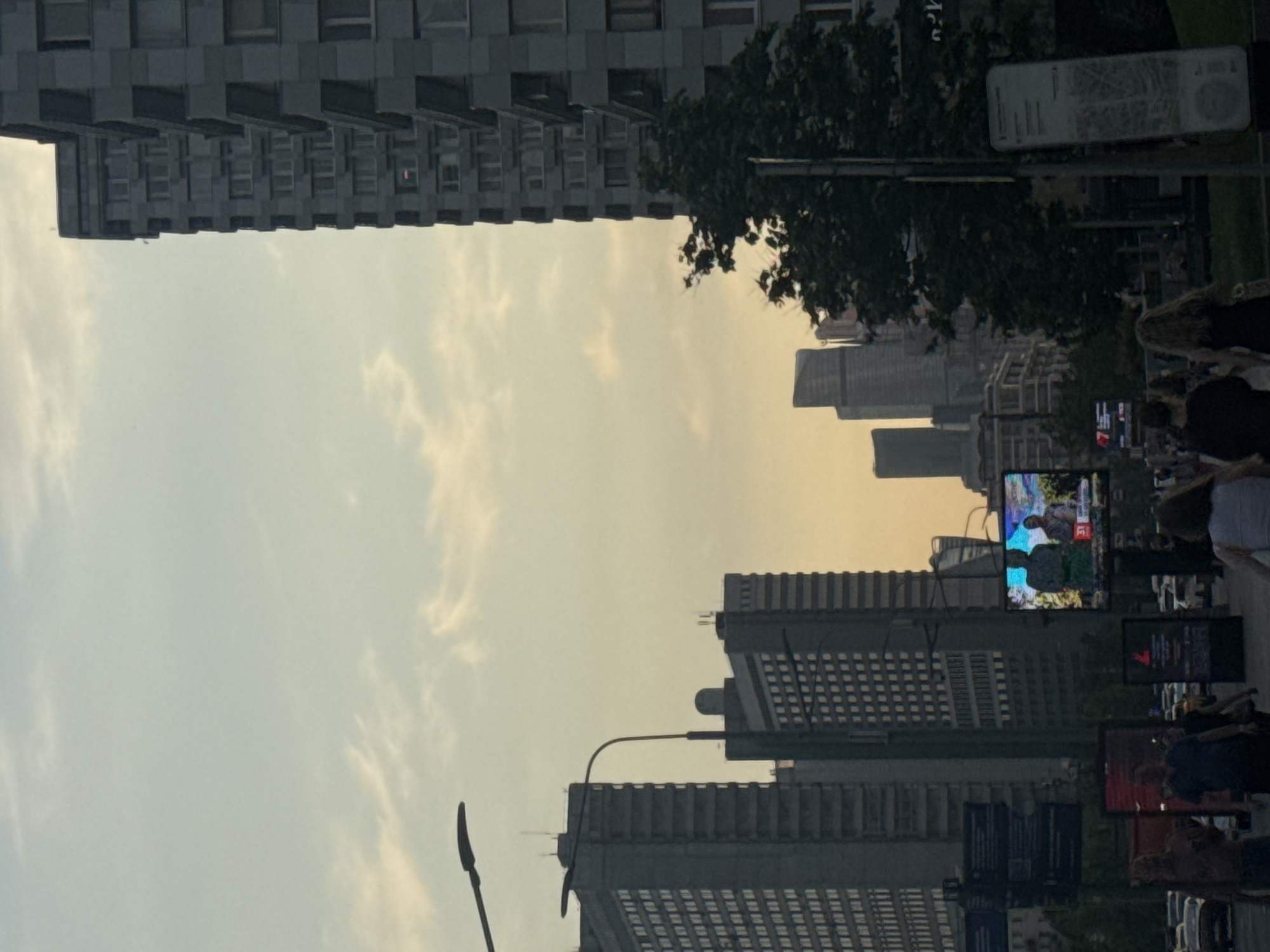 The image depicts a street scene in Moscow, Russia. Tall buildings dominate the view, with a large, gray, Soviet-era apartment block on the top left and more modern skyscrapers in the distance on the right. People are walking on the street, partially obscured by billboards and other street furniture. A video screen displays content, showing figures and shapes. The sky is hazy, with a yellow-orange tint, suggesting sunset or a polluted atmosphere. Streetlights and foliage are interspersed throughout the scene.