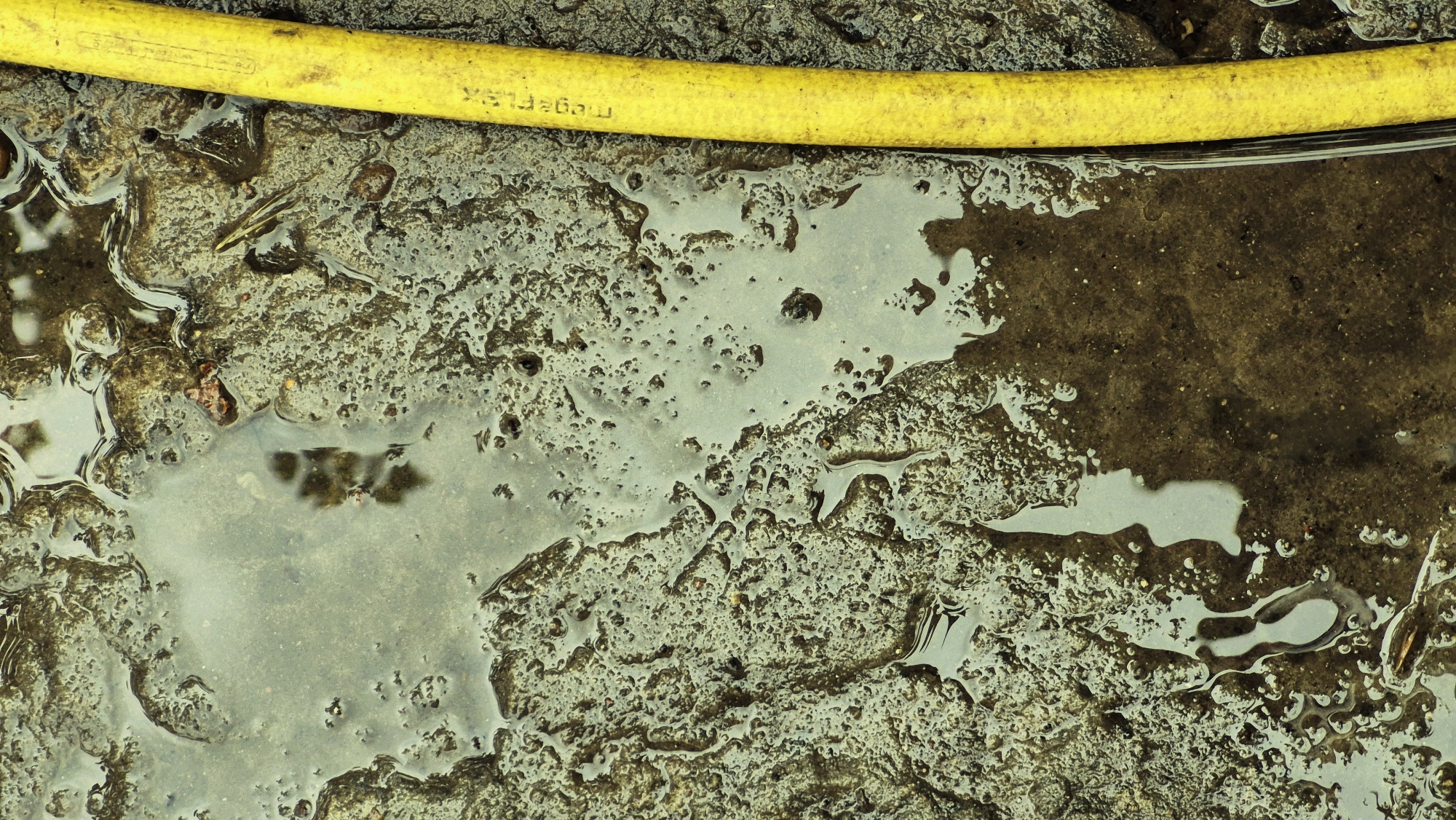 The image is an extreme close-up, high-angle shot of a wet, muddy, and textured surface with a bright yellow hose running vertically along the right side. The ground appears to be a mixture of soil, small stones, and water, with numerous small indentations and bubbles on the surface of the mud. Reflections of light are visible on the wet areas, creating variations in tone and texture. The yellow hose has some text printed on it, which appears to be "megaflex". The overall impression is of a somewhat gritty and weathered outdoor environment, possibly in a backyard or construction area. The specific location in Ialoveni, Moldova, is not directly ascertainable from the visual elements of the image itself.