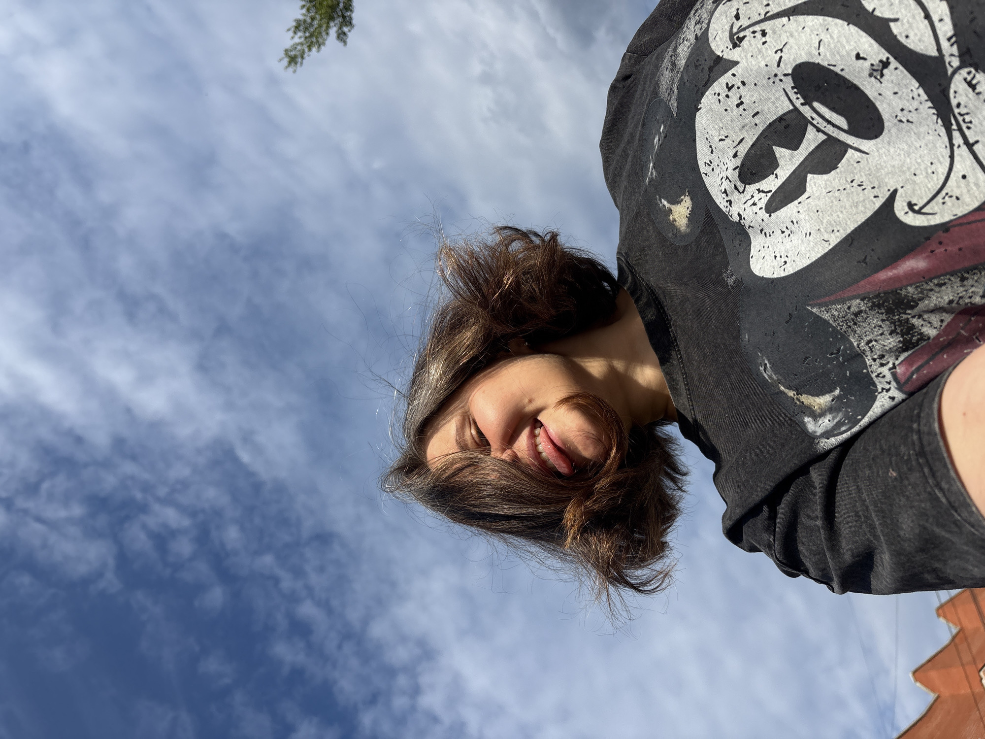 The media file is a close-up, low-angle shot of a young person looking up towards the sky. The subject has brown hair that is partially covering their face, and they are smiling with their tongue slightly sticking out. They are wearing a dark t-shirt with a distressed graphic print of a cartoon character, possibly Mickey Mouse. The background is a bright blue sky with scattered white clouds, suggesting it is daytime. There is a hint of greenery from a tree branch in the upper left corner, and a portion of an orange building is visible in the lower right corner. The lighting indicates a sunny day. The image appears to be a selfie taken outdoors.