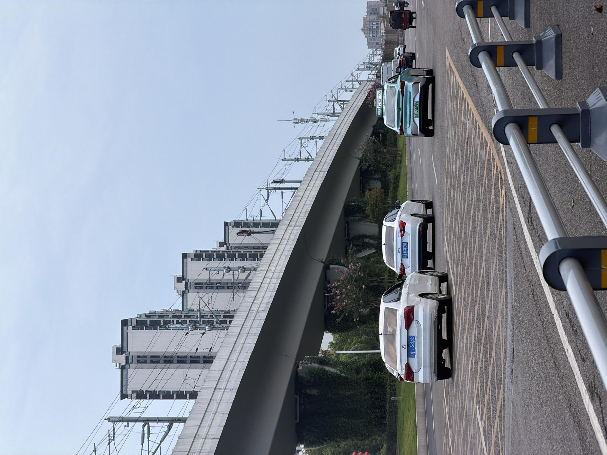 In Jiangjin, China, a multilane road passes under an elevated structure, likely a railway or highway, supported by concrete pillars. Overhead, complex electrical lines for transportation are visible. Several vehicles are traveling on the road, including white sedans, a teal electric car, and a three-wheeled vehicle. Buildings, some with modern architecture and many windows, are situated in the background. Greenery, including vines climbing the pillars and trees with pink blossoms, adds a natural element to the urban environment. The sky is overcast and pale blue. The scene depicts typical urban transportation infrastructure and activity in the region.