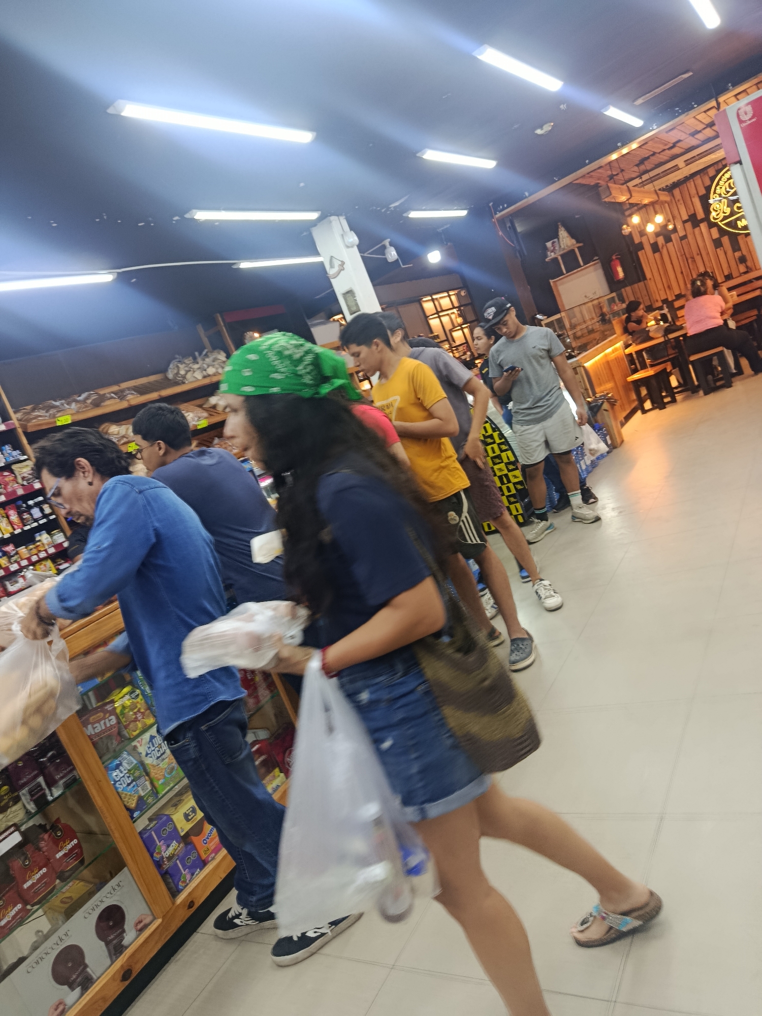 Here is a detailed description of the image:
**Scene:** The image appears to be taken inside a grocery store or bakery in Altagracia, Venezuela. The location appears to be well-lit with overhead fluorescent lights and decorated with rustic, wooden paneling and lighting fixtures.
**People:** There are several people in the frame, standing in a queue. The focus is on a line of customers who are likely waiting to pay for their purchases. The closest person to the camera is a woman wearing denim shorts, a dark blue shirt, a green bandana, carrying a tote bag, and holding a plastic bag with what looks like bread in it. Behind her is a line of people of varying ages and attire, including people wearing shorts, t-shirts, and hats.
**Subjects/Objects:** The foreground includes shelves stocked with various food items such as snacks, cookies, and coffee products. The customers are holding or carrying plastic bags, presumably filled with their purchased items. In the background, there is a counter area where people are sitting and eating.
**Actions:** The primary action occurring is customers waiting in line. Some customers in the background are seated at tables, eating.
**Interactions:** The image seems to capture a normal scene of commerce, where people are selecting and purchasing goods and enjoying the atmosphere.
**Environment:** The setting is a public space, most likely a grocery store or bakery with a small cafe area. The store seems clean and well-maintained, with a generally relaxed atmosphere.