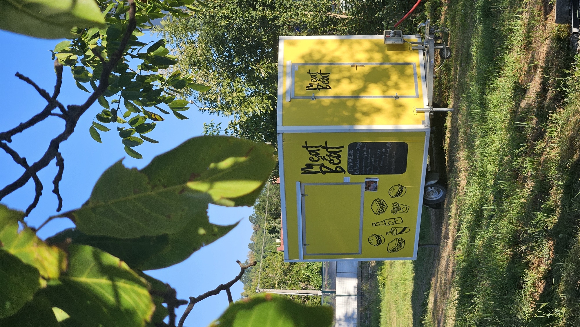 A bright yellow food truck, branded "Meat Beat", is parked on a grassy area under a clear blue sky. The truck features a serving window and a door, both with the "Meat Beat" logo. A menu is displayed on the side of the truck, featuring illustrations of burgers, fries, and drinks, along with text in a language that appears to be Slovak. The truck is surrounded by lush green grass and mature trees. In the background, there is a white fence and more trees, with a hint of a building and hills in the distance. The overall scene suggests an outdoor food vendor operating in a somewhat rural or suburban setting. The image was captured in the city of Prešov, Slovakia.