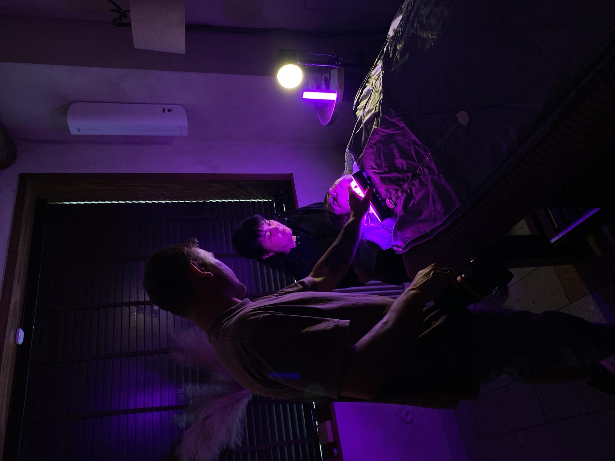 The image depicts an indoor scene illuminated by purple and yellow lights. A person is lying on a massage table, covered by a dark material. Another person, wearing a dark top, is standing over them, holding a rectangular device emitting purple light towards the person on the table. A third person, wearing a light-colored t-shirt, is standing to the left, holding a black device. In the background, there are dark blinds, a white vertical device mounted on the wall, and a lamp on a small table. The overall impression is of a spa or wellness treatment session. The location is not specified as Izhevsk, Russia.