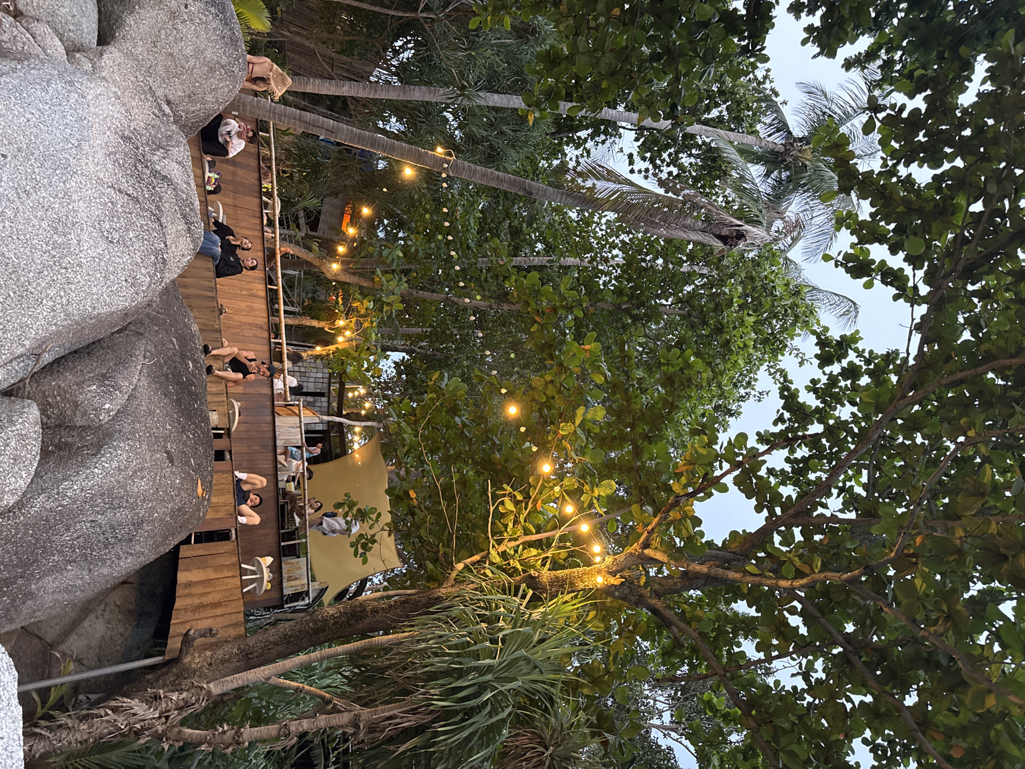 In Karon, Thailand, a group of people are gathered at an outdoor establishment situated amidst large rocks and lush tropical foliage. The establishment features wooden platforms and railings, illuminated by a string of warm, incandescent lights. Several individuals are seated and reclining on these platforms, appearing relaxed and engaged in conversation or observation. One person, wearing a straw hat, is positioned on a prominent rock in the foreground. The scene is set against a backdrop of palm trees and dense green trees, suggesting a beachside or coastal environment. The lighting indicates it is either dusk or dawn.