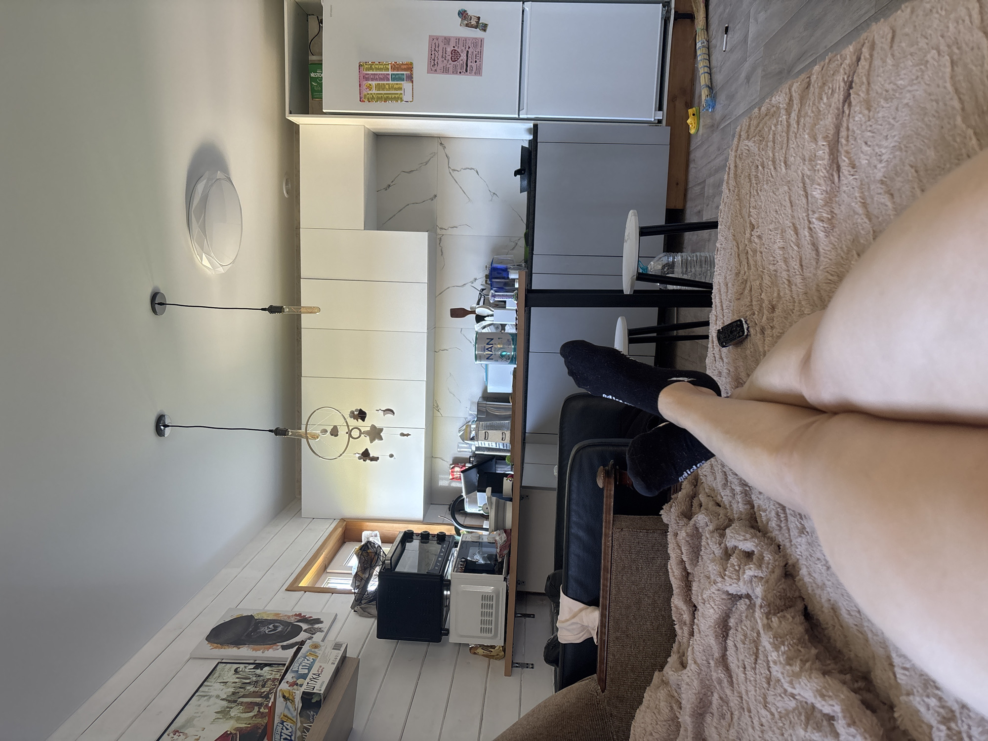 The image shows a person lying on a couch in an indoor setting, possibly an apartment, near Раздолье, Russia. The person is wearing black socks and has their legs crossed. A beige blanket is on the couch. In the background is a kitchen with white cabinets and appliances, including a refrigerator and a microwave on the counter. There is also a dining table with chairs. Light fixtures and artwork adorn the walls. The environment appears to be a residential living space.