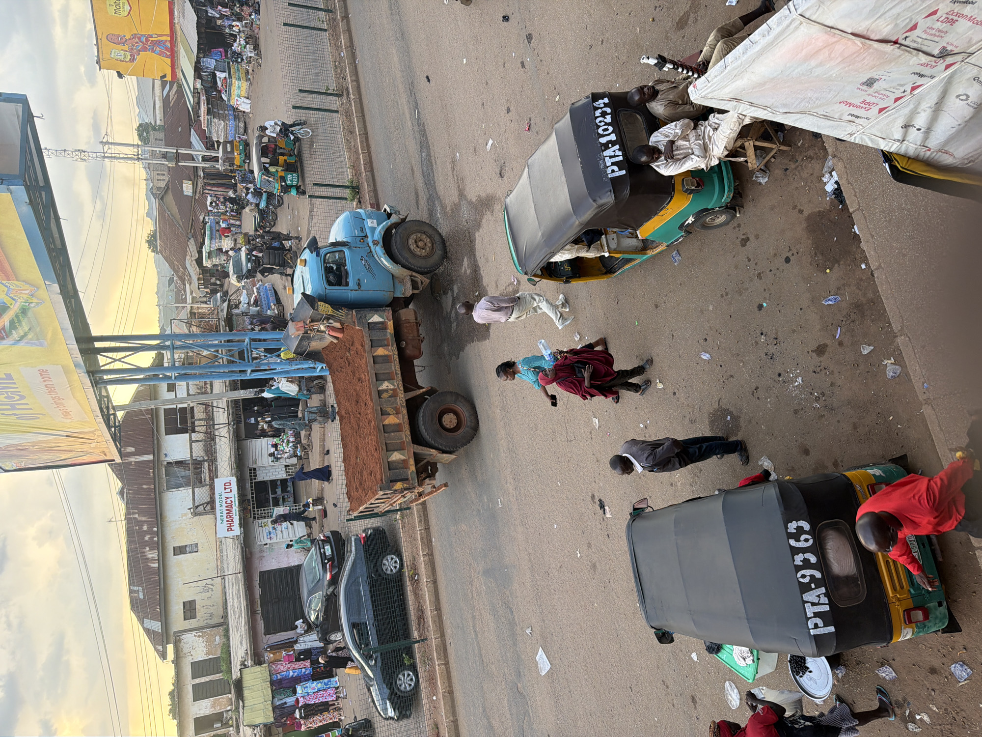 The image depicts a street scene in Bukuru, Nigeria, with a mix of commercial activity and pedestrian traffic. A light blue dump truck filled with red soil is parked in the foreground, partially obscuring a black car. Several people are walking on the street, some carrying items. A green and yellow auto-rickshaw with the registration "PTA-9363" is stopped, with several individuals around it. Another auto-rickshaw, "PTA-10234", is also visible. Buildings line the street, some with corrugated metal roofs, and stalls are set up selling various goods, including fabrics and what appear to be mattresses. The sky is overcast with some hints of sunlight. The overall impression is a bustling marketplace environment.