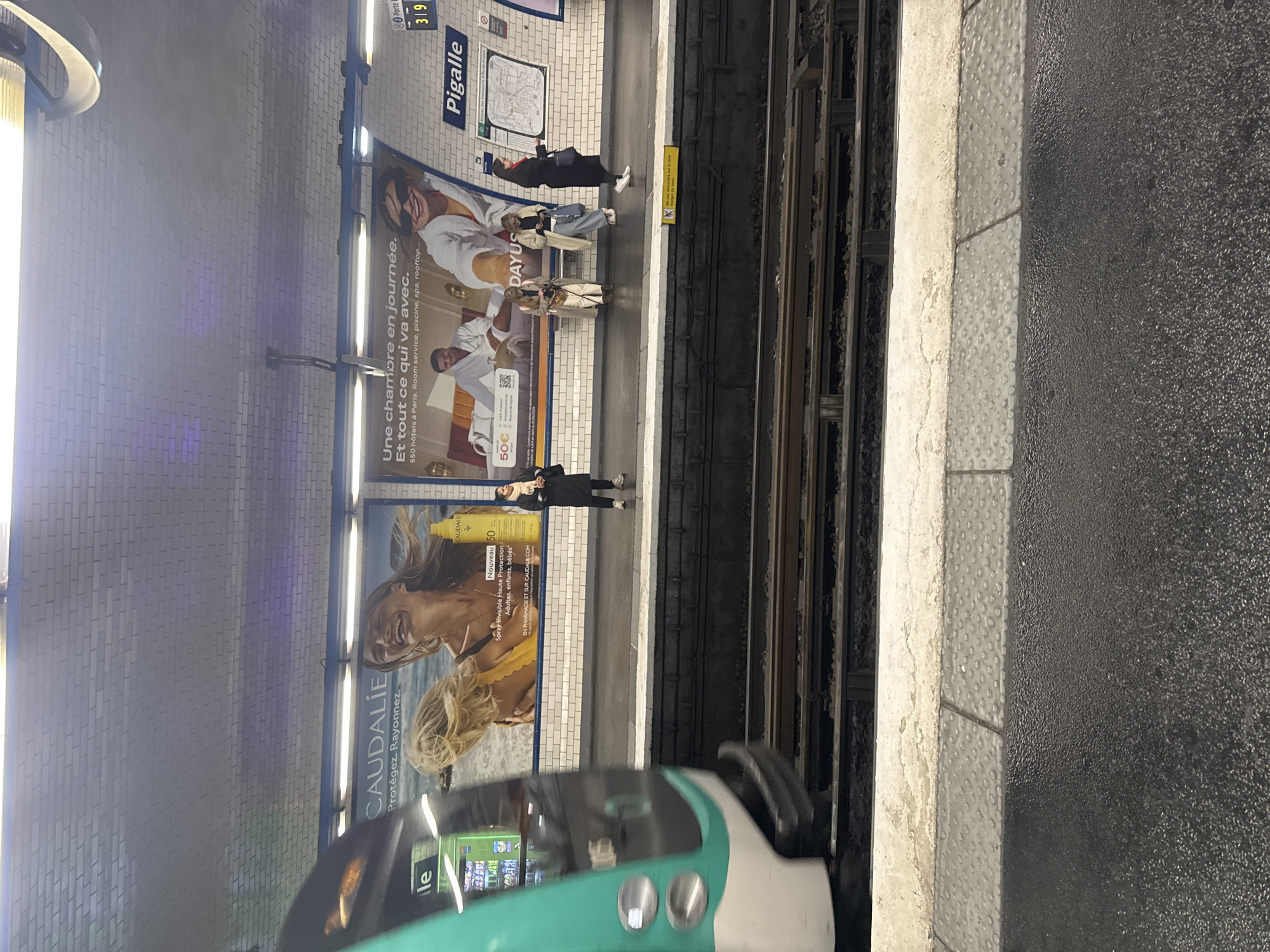 The image captures a scene inside the Pigalle metro station in Paris, France. Several people are waiting on the platform, standing near advertising posters for Caudalie and Dayus. A green and white metro train is pulling into the station. The platform edge is marked by a textured gray strip. Above, the station name 'Pigalle' is visible on a sign along with route information.