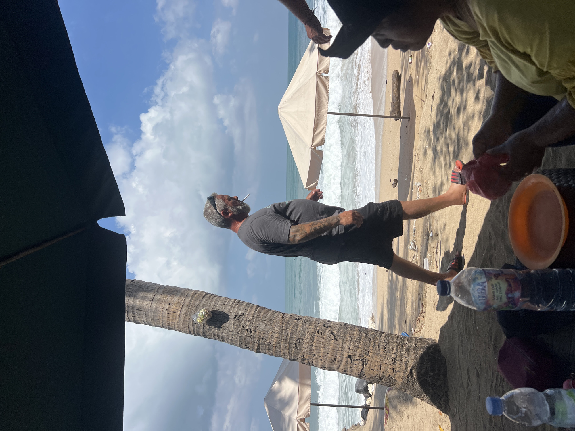A man with a gray beard, tattoos on his arms, and wearing sunglasses, a dark t-shirt, and black shorts walks along the beach in Kuta, Indonesia, smoking a cigarette. He is walking past a palm tree and an umbrella. In the foreground, two people are seated under an umbrella, with bottles of water and an orange bowl on a table. The ocean with waves is visible in the background, along with other umbrellas and a clear blue sky with clouds.