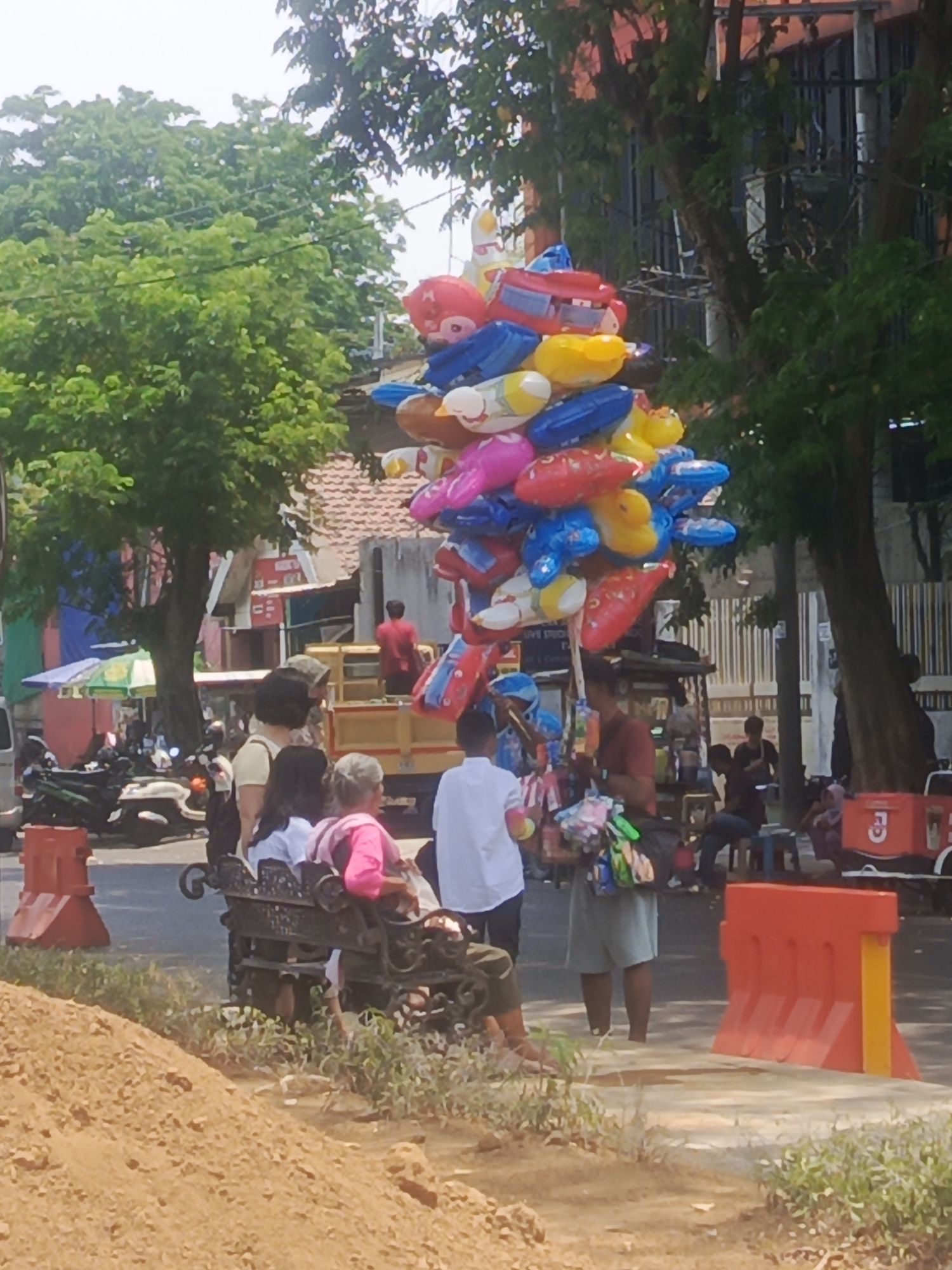 A street scene in Surabaya, Indonesia shows a vendor selling a large bunch of inflatable balloons from a cart. The balloons are shaped like cartoon characters and animals. The vendor, a man wearing a red shirt and light-colored shorts, is standing behind the cart. Several people are visible in the background, including a group of women and children sitting on a bench, a man standing near a truck, and other individuals walking or standing. The environment appears to be a busy street with buildings, trees, and parked motorcycles. Orange traffic barriers are placed along the side of the road. The overall impression is a typical street scene with commercial activity in an urban Indonesian setting.