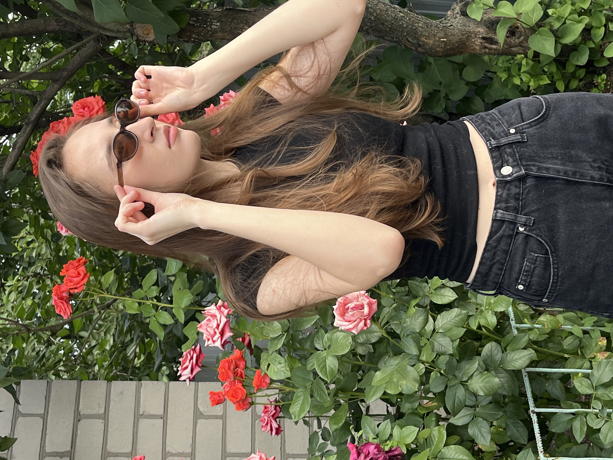 A young woman with long brown hair stands outdoors in front of a brick wall and a rose bush. She is wearing a black t-shirt and black jeans and sunglasses. She is holding her sunglasses up to her face with both hands. The background consists of green foliage and a tree trunk.