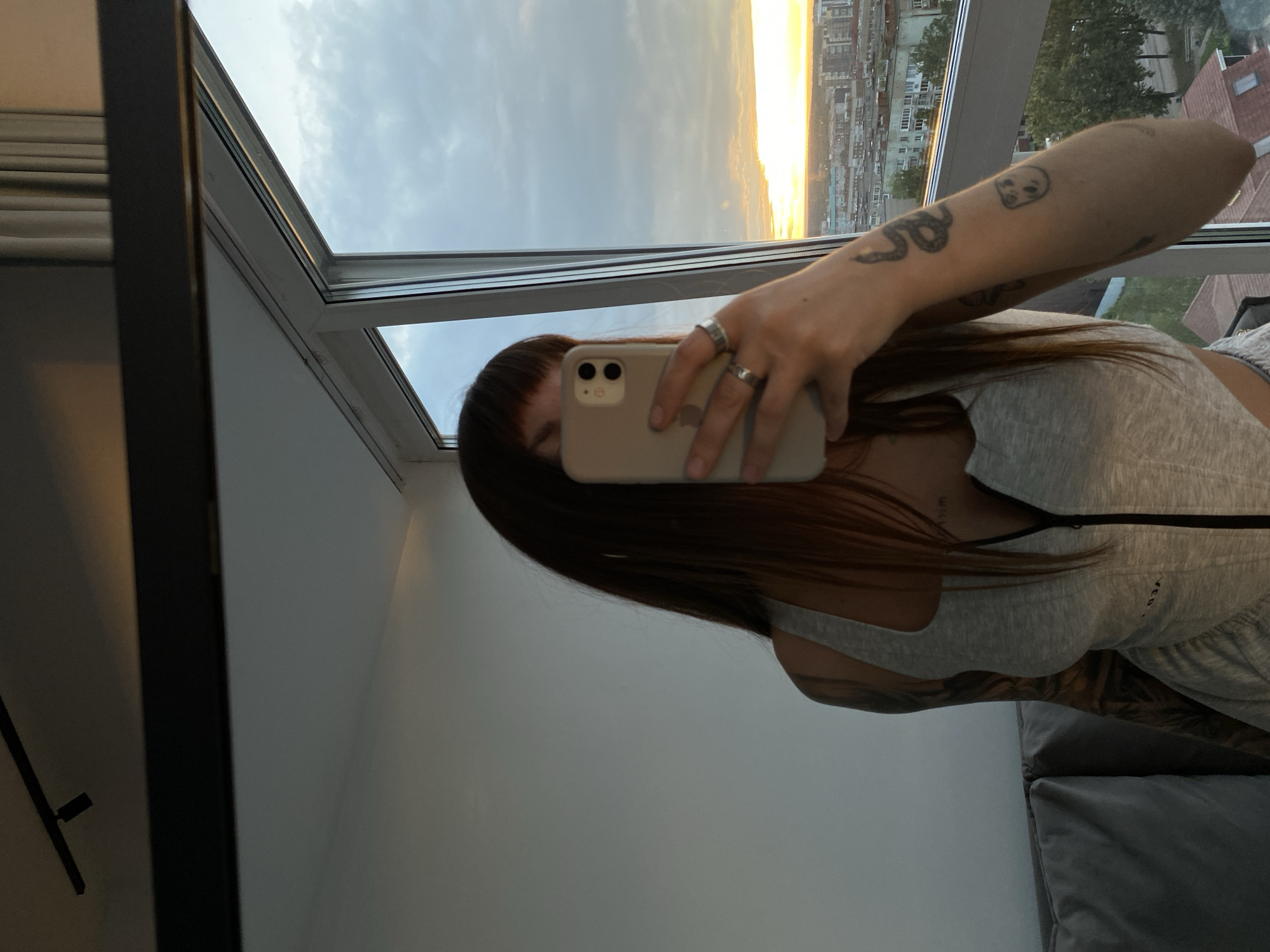 A person is taking a selfie in a room with a large window overlooking a city. The person's face is partially obscured by their phone, which they are holding up to take the picture. They have long, dark hair and are wearing a grey tank top. There are tattoos on their arm, including a snake and a small skull. The person is wearing rings on their fingers.
The window shows a sky with clouds and a bright yellow sunset. Buildings are visible in the distance, suggesting an urban setting. The room appears to be modern, with a large black frame around the window and white walls. A dark grey sofa is visible in the lower right corner. The lighting suggests it is either late afternoon or early evening.