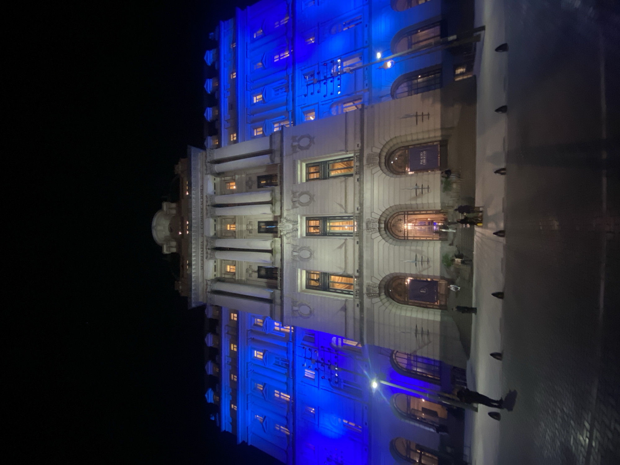 The image shows a building at night, illuminated with blue lighting on the upper floors. The lower floors have a more muted, warmer lighting from within. The architecture appears to be classical or neoclassical, with arched windows and decorative features. People are standing near the entrance. The image is rotated, so the sky is on the left and the ground is on the right. The building is likely the Palacio de Correos, also known as the Centro Cultural Kirchner, located in Buenos Aires, Argentina.