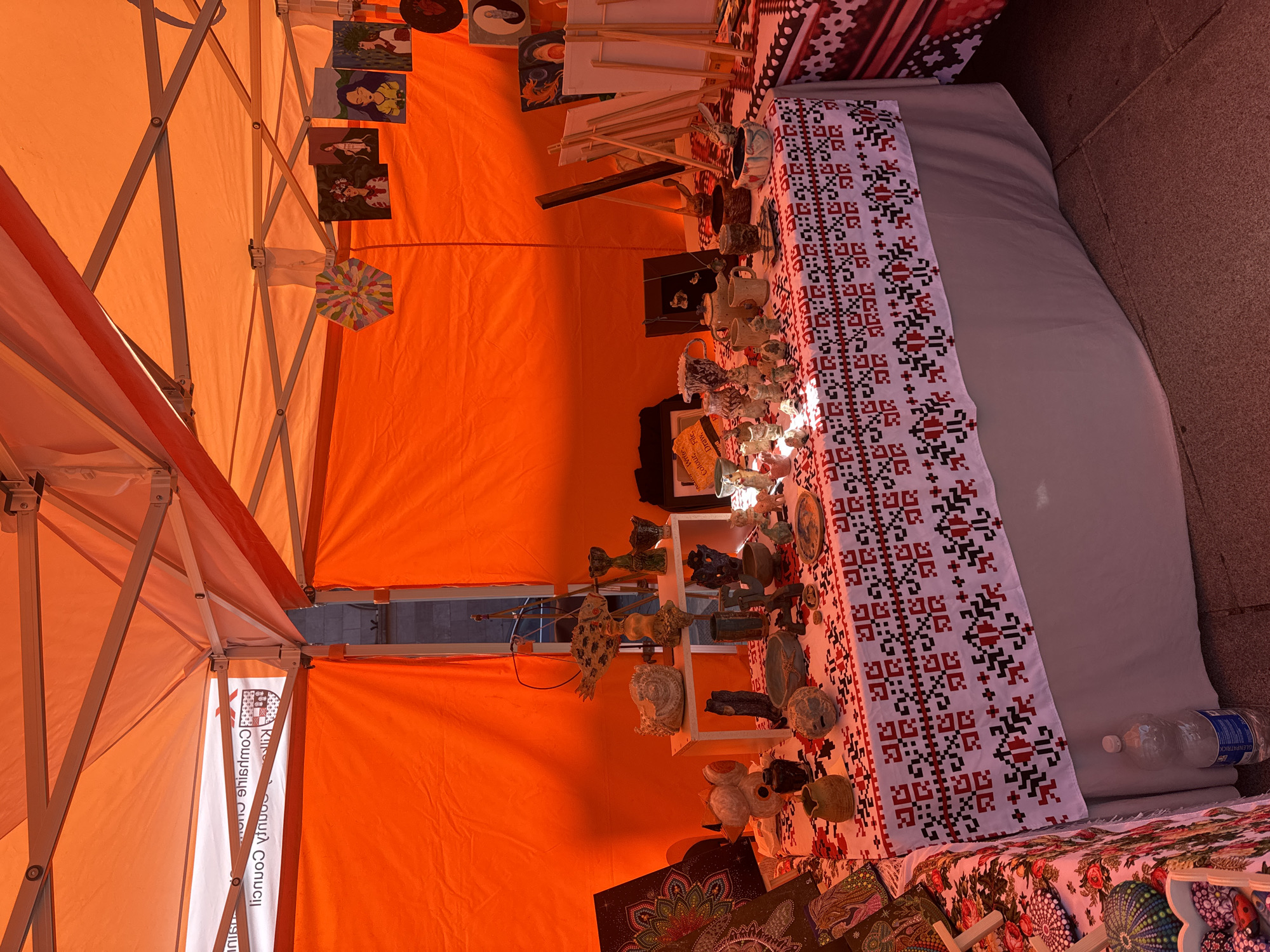 An outdoor market stall in Kilkenny, Ireland, is covered by an orange canopy. The stall displays a variety of handcrafted items, including pottery, small figurines, and artwork. A table is draped with a white cloth featuring a red and black folk pattern. On the table, there are numerous ceramic pieces, some appearing to be animals and teapots, along with other decorative objects. To the left, an easel holds a canvas with a mandala-like design. In the background, several small paintings of people are hung on the stall's fabric wall. The scene suggests a local craft fair or market.