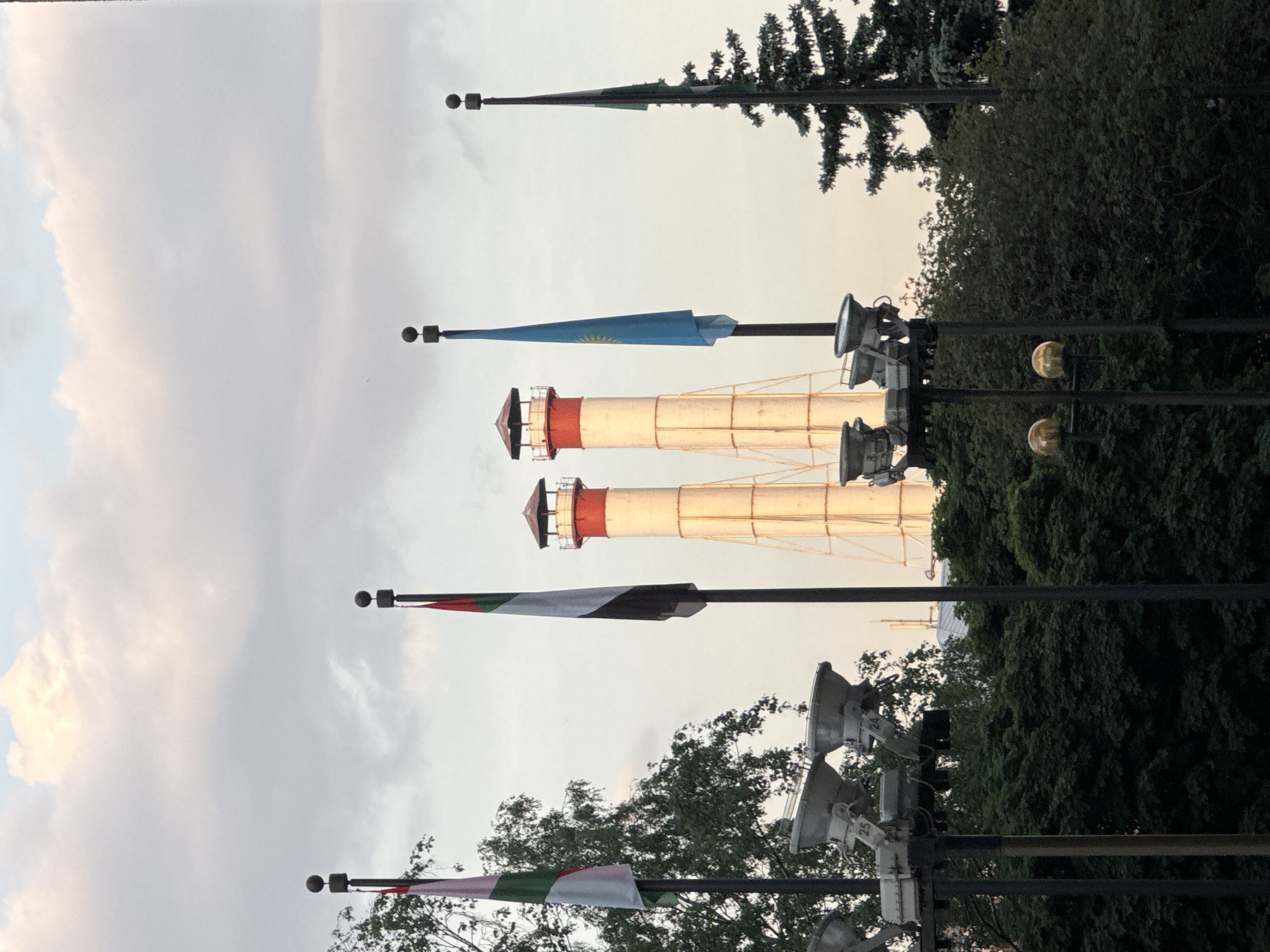 The image shows a view of the Vodovzvodnaya Tower, also known as Water Tower, and flags in Moscow, Russia. The tower has a white cylindrical body with red and white horizontal bands and a red rooftop. Several flags are positioned on poles, and they are partially obscured by the surrounding trees and streetlights. The sky is overcast.