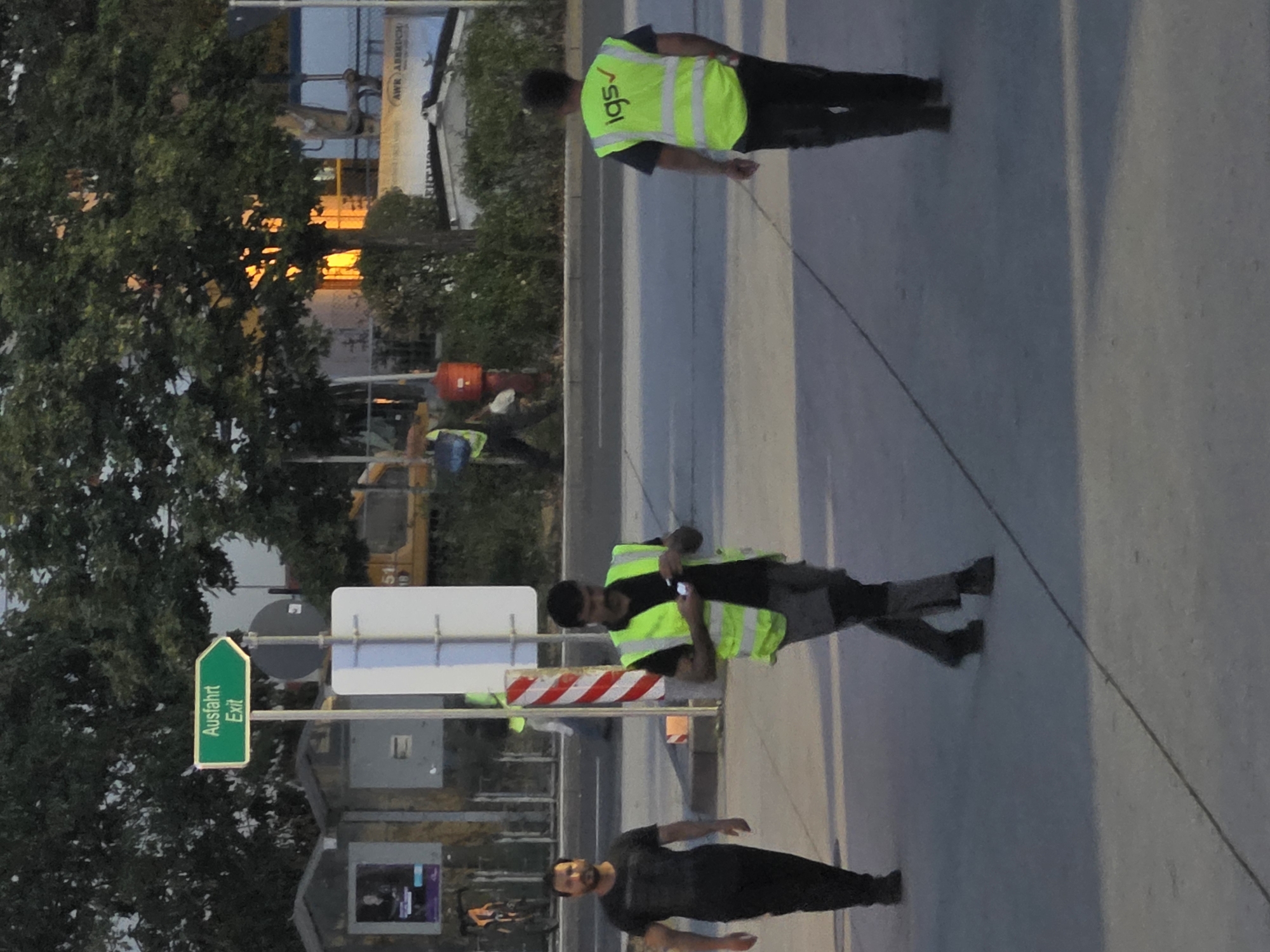 The image depicts an outdoor scene with three men walking on a paved area. In the background, trees are visible, along with a yellow construction vehicle and a sign that reads "Ausfahrt Exit." The man on the left is wearing a black t-shirt and dark pants. The man in the middle is wearing a high-visibility yellow vest over a dark shirt and light-colored pants, and is holding a mobile phone. The man on the right is also wearing a high-visibility yellow vest with the logo "igs ✓" and dark pants. The lighting suggests it might be dusk or early morning. There are no specific landmarks in the image that definitively place it in Frankfurt, Germany.