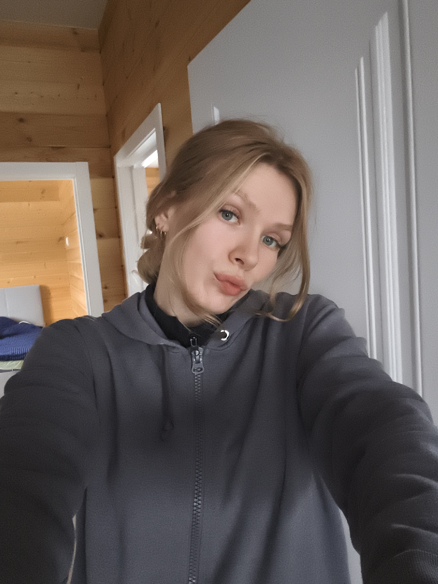 The image shows a fair-skinned young woman with light blonde hair and blue eyes, wearing a gray zip-up hoodie over a black top. Her hair is partially pulled back, revealing small hoop earrings. She is making a pouting expression. The background features a light-colored wooden wall on the left and a white paneled door on the right. In the background to the left, a doorway leads into another room with a bed visible. There is no information in the photo to verify the location.