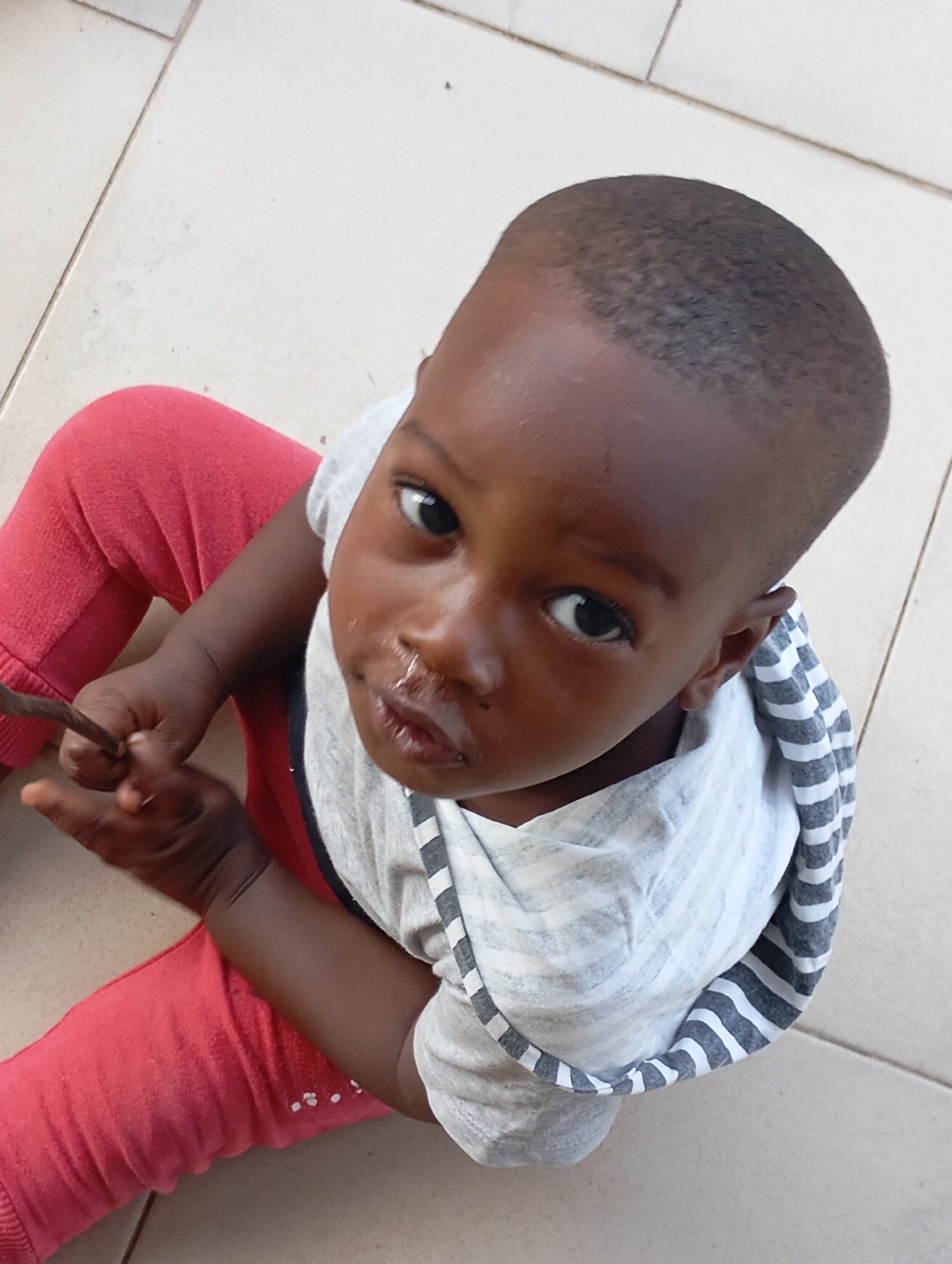 A young, dark-skinned child sits on a light-colored tile floor, possibly in a domestic setting in or near Barikin Jirga Summa, Nigeria. The child wears red pants and a gray and white striped top. The child looks upward at the camera and holds a small, dark twig in their hands. The child has a runny nose.