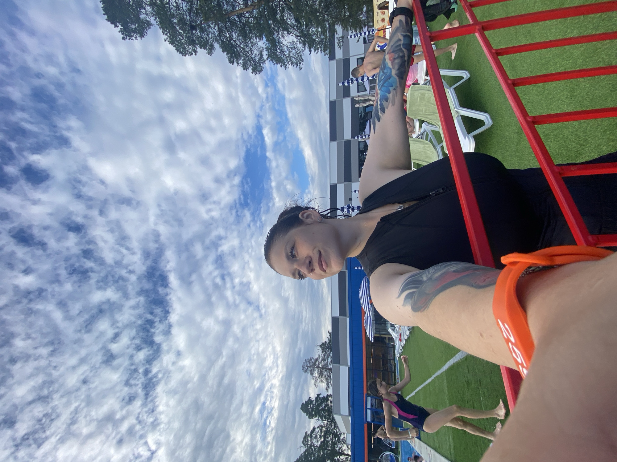 A woman with dark hair and tattoos on her arms is taking a selfie in an outdoor area. She is wearing a black sleeveless top and an orange wristband. In the background, a young girl in a swimsuit is running on a green surface, and there are lounge chairs and umbrellas. The sky is cloudy. The image appears to be taken at the Votkinsk Recreation Complex, located in the city of Votkinsk, Russia, which is known for its water park and other recreational facilities.