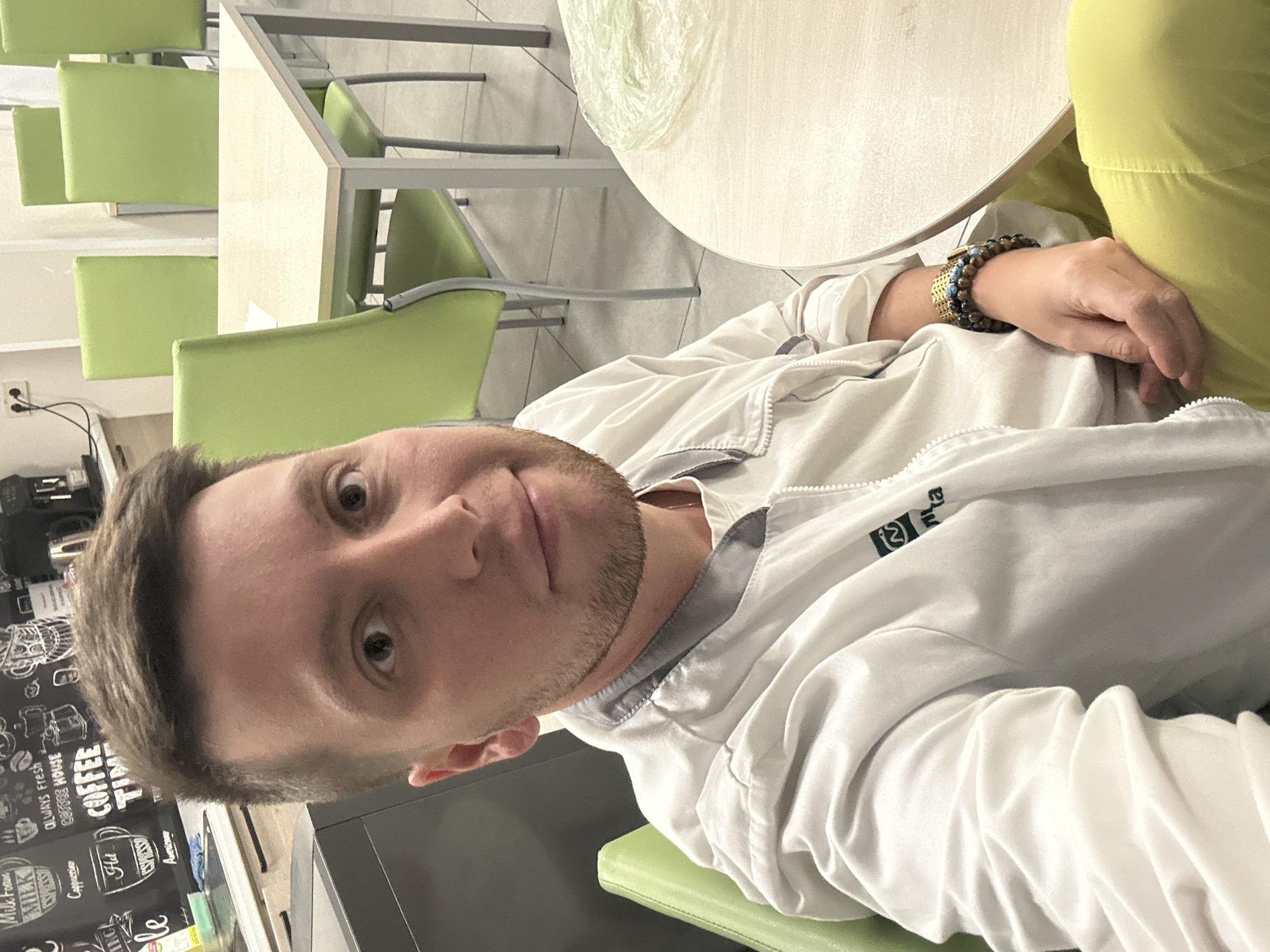 A selfie was taken indoors in a casual environment. The subject is a man with short brown hair and stubble wearing a white jacket. He is seated in a green chair. A round table is to the left. In the background, there are other green chairs and tables. A sign with coffee-related information, including "COFFEE TIME" and "ESPRESSO," is visible. The location may be a cafe or breakroom. The city is not specified.